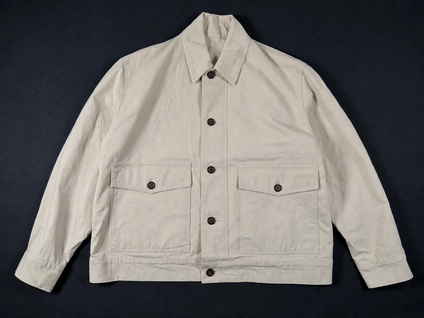 An unbranded ecru chore jacket laid flat on a near-black surface, lit evenly from above.
