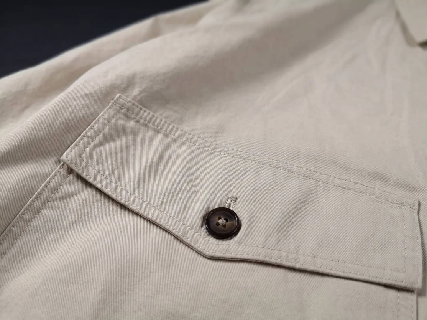 A close view of the same ecru jacket's chest pocket: the flap's double topstitching and a brown four-hole button.