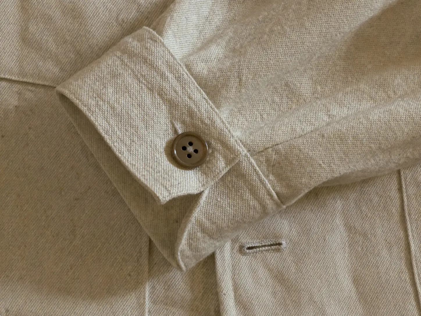 An extreme close-up of the same ecru chore jacket's cuff: the folded sleeve edge, its topstitching, and a fastened brown four-hole button.