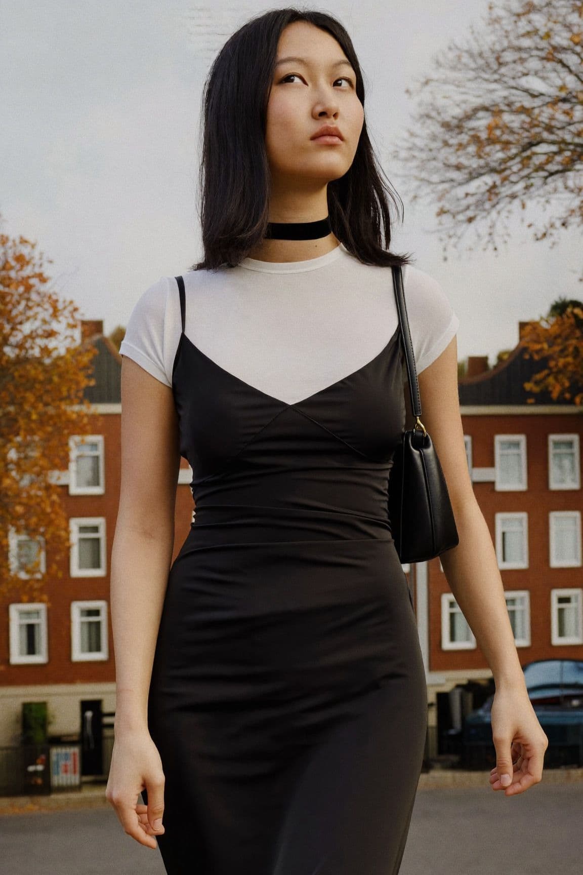 A woman in a black satin slip dress layered over a white t-shirt with a thin black choker on a street of brick houses