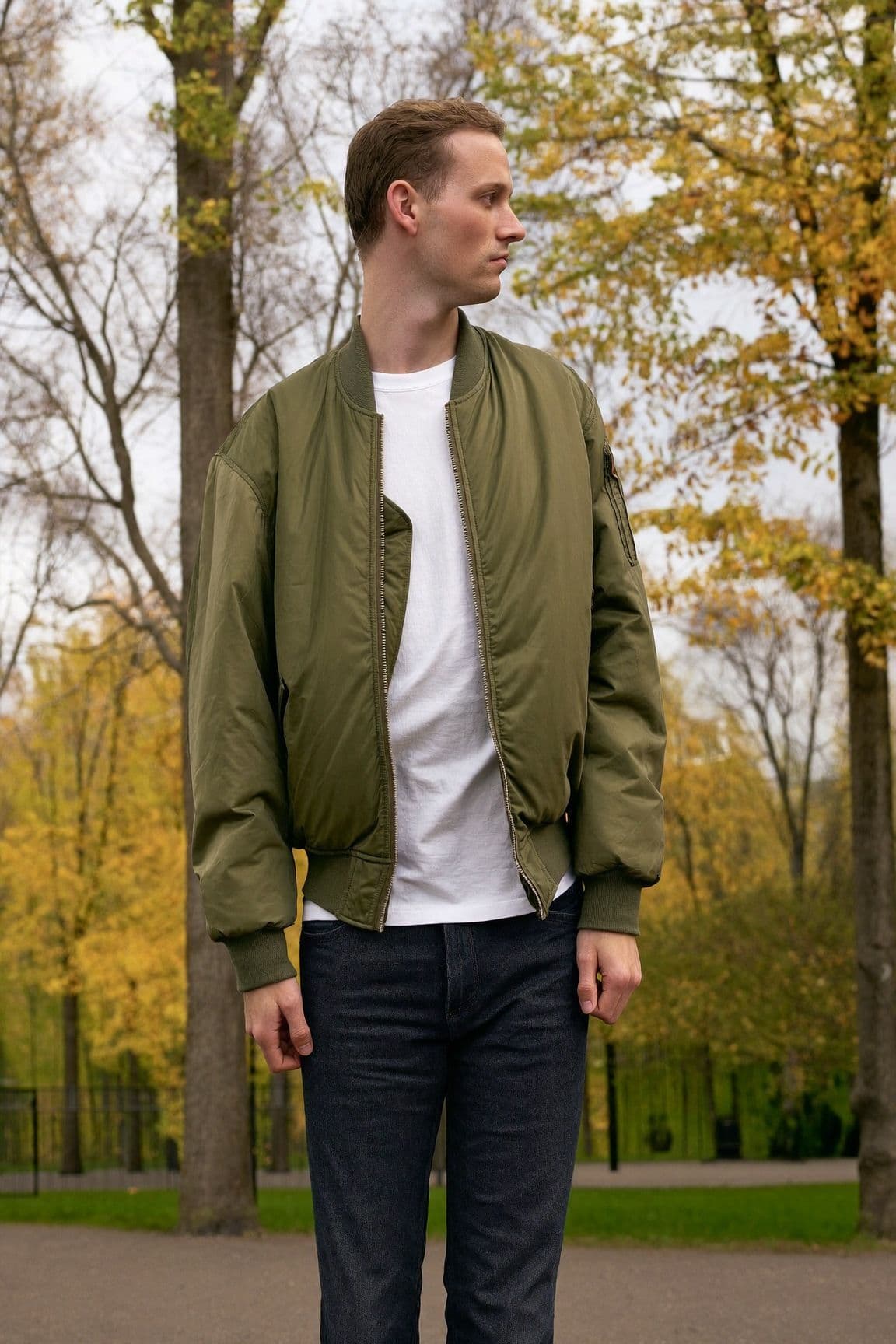 A man in an olive bomber jacket over a white t-shirt and dark slim jeans standing in a park in autumn