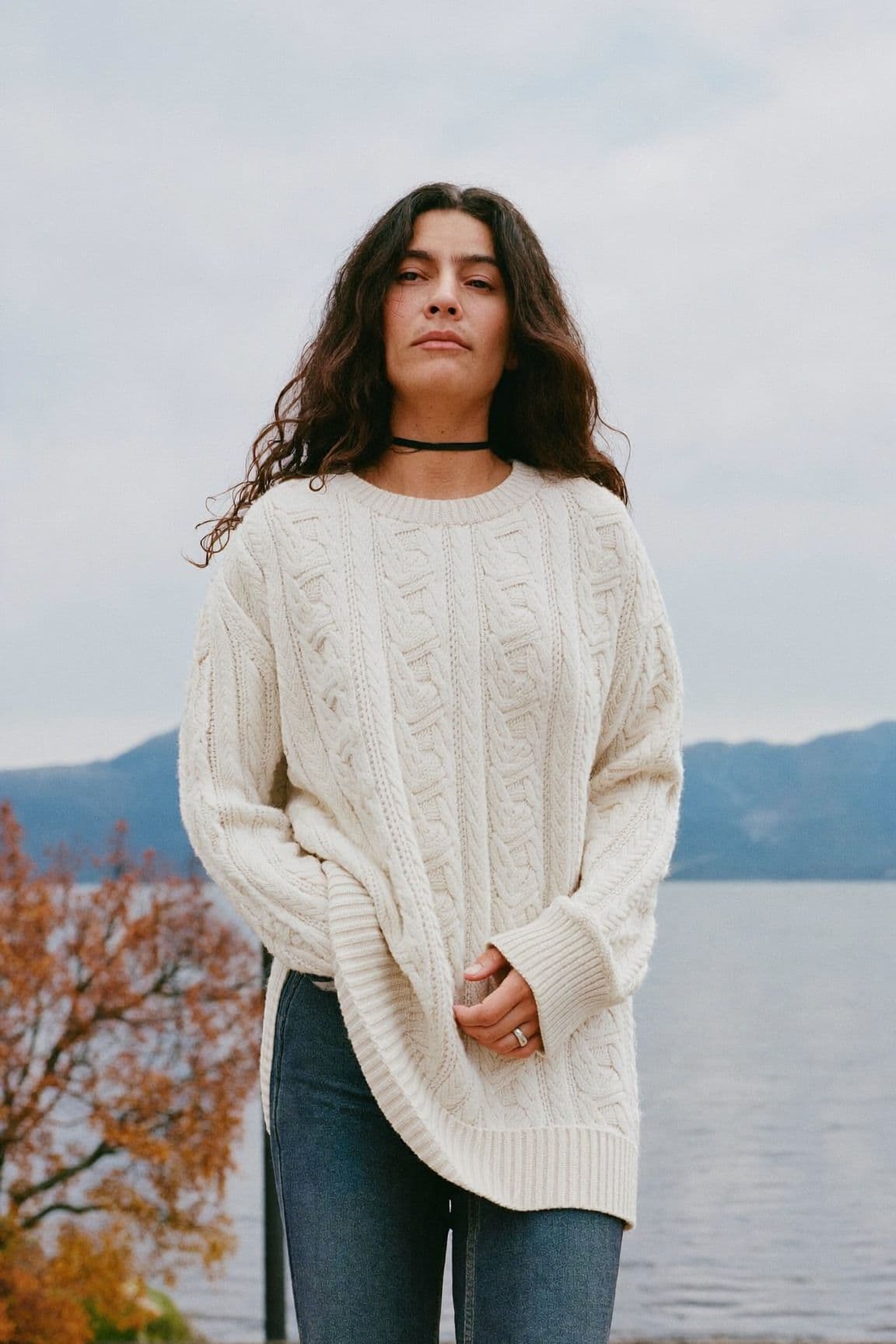 A woman in an oversized cream cable-knit sweater, skinny jeans and a black choker by the water
