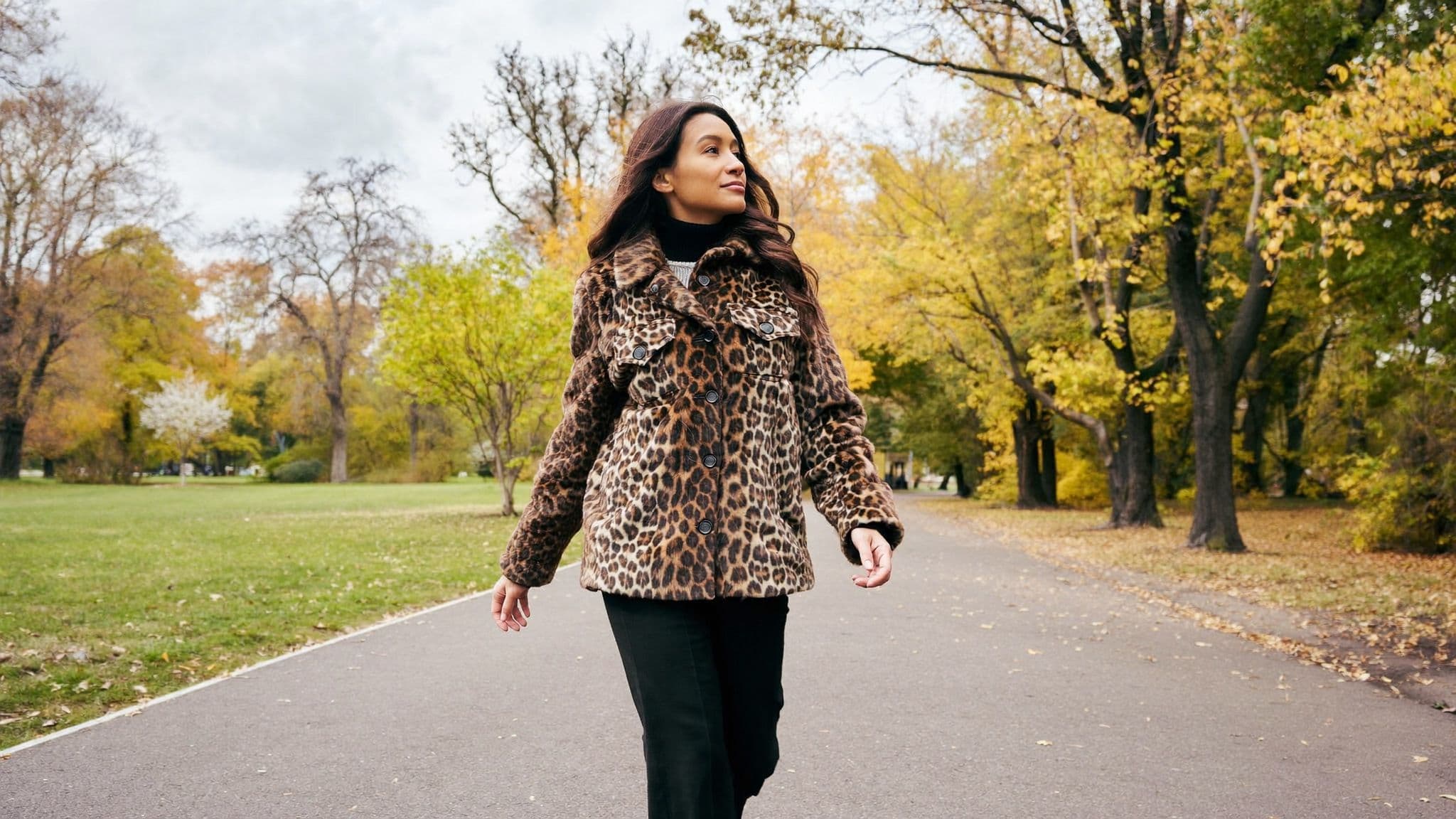 A woman in a leopard-print faux-fur jacket over a black turtleneck and black trousers walking along a park path