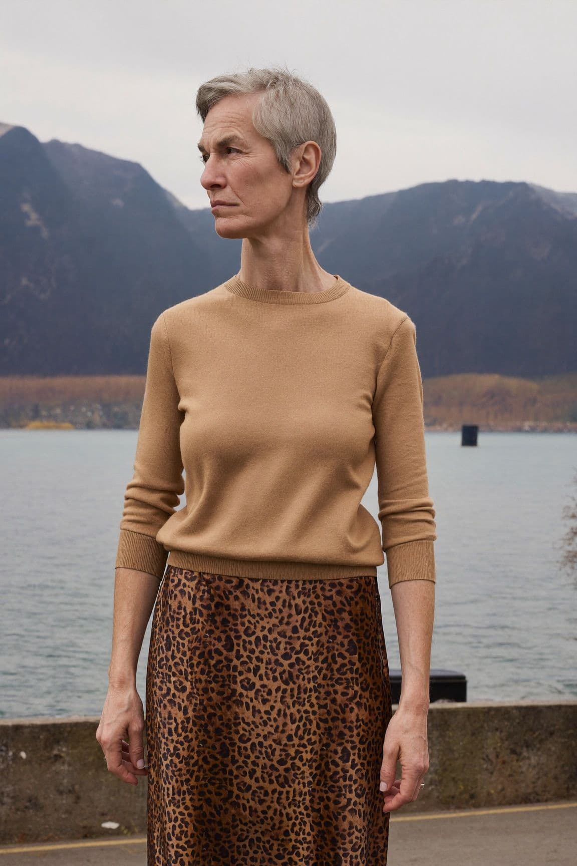 A woman with short grey hair in a camel sweater and a leopard-print satin midi skirt by the water