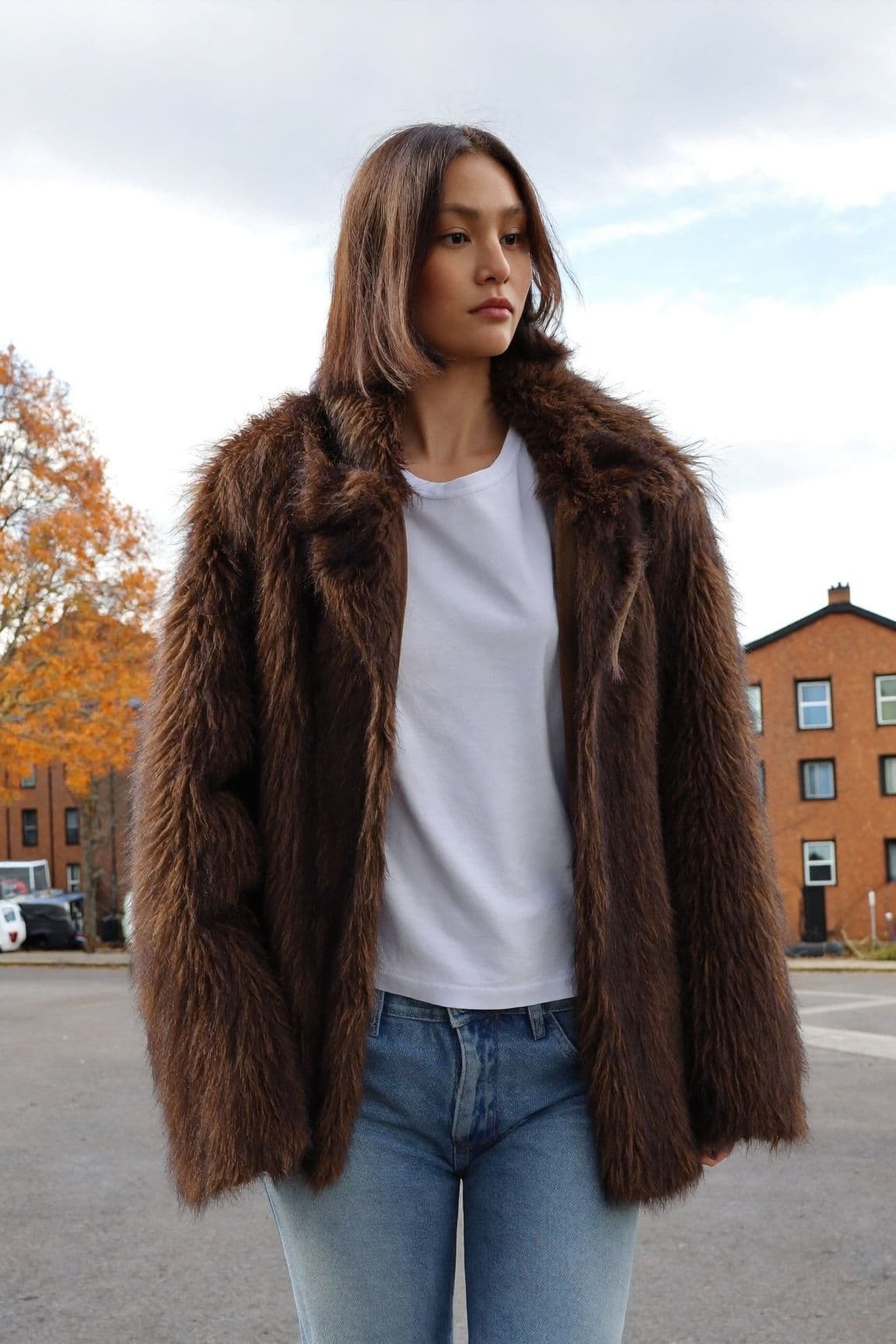 A woman in a chocolate-brown faux-fur coat over a white t-shirt and light-blue jeans on a residential street