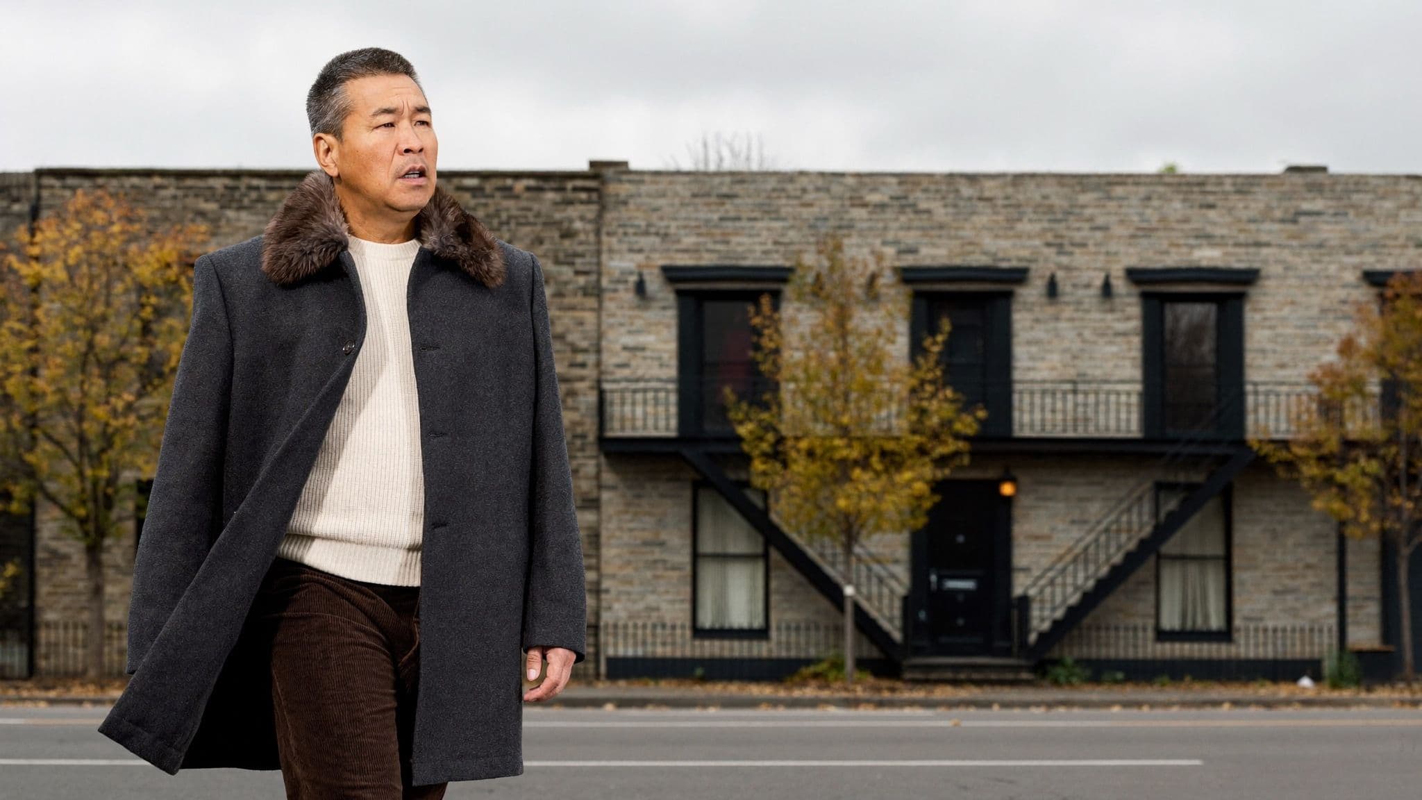 Man walking past a stone building in Montreal in autumn, wearing a charcoal wool overcoat with a brown faux-fur collar, a cream ribbed sweater and brown corduroy trousers