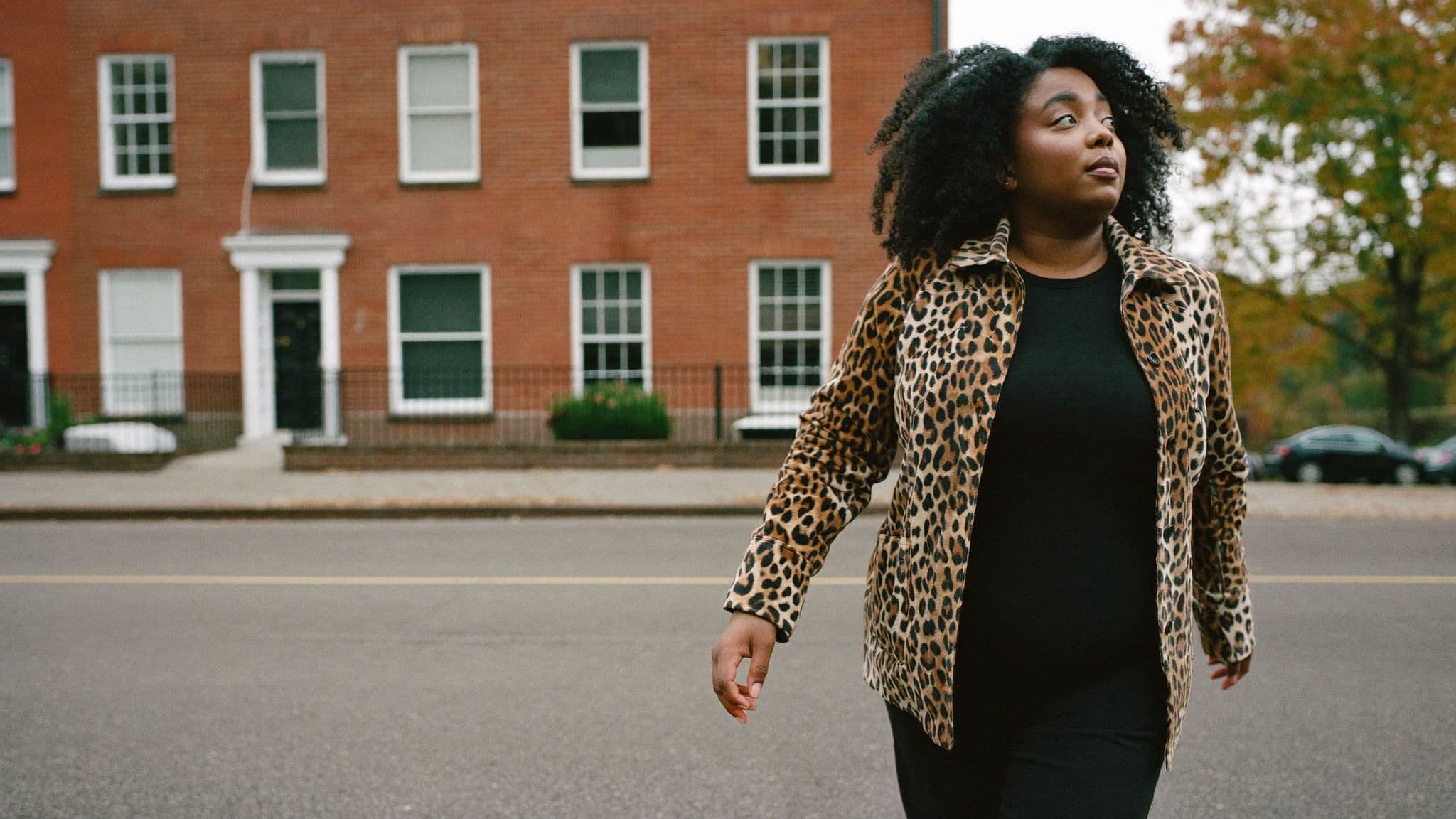 Woman walking on a Toronto street lined with red-brick houses, wearing a leopard-print jacket open over a plain black top and black trousers