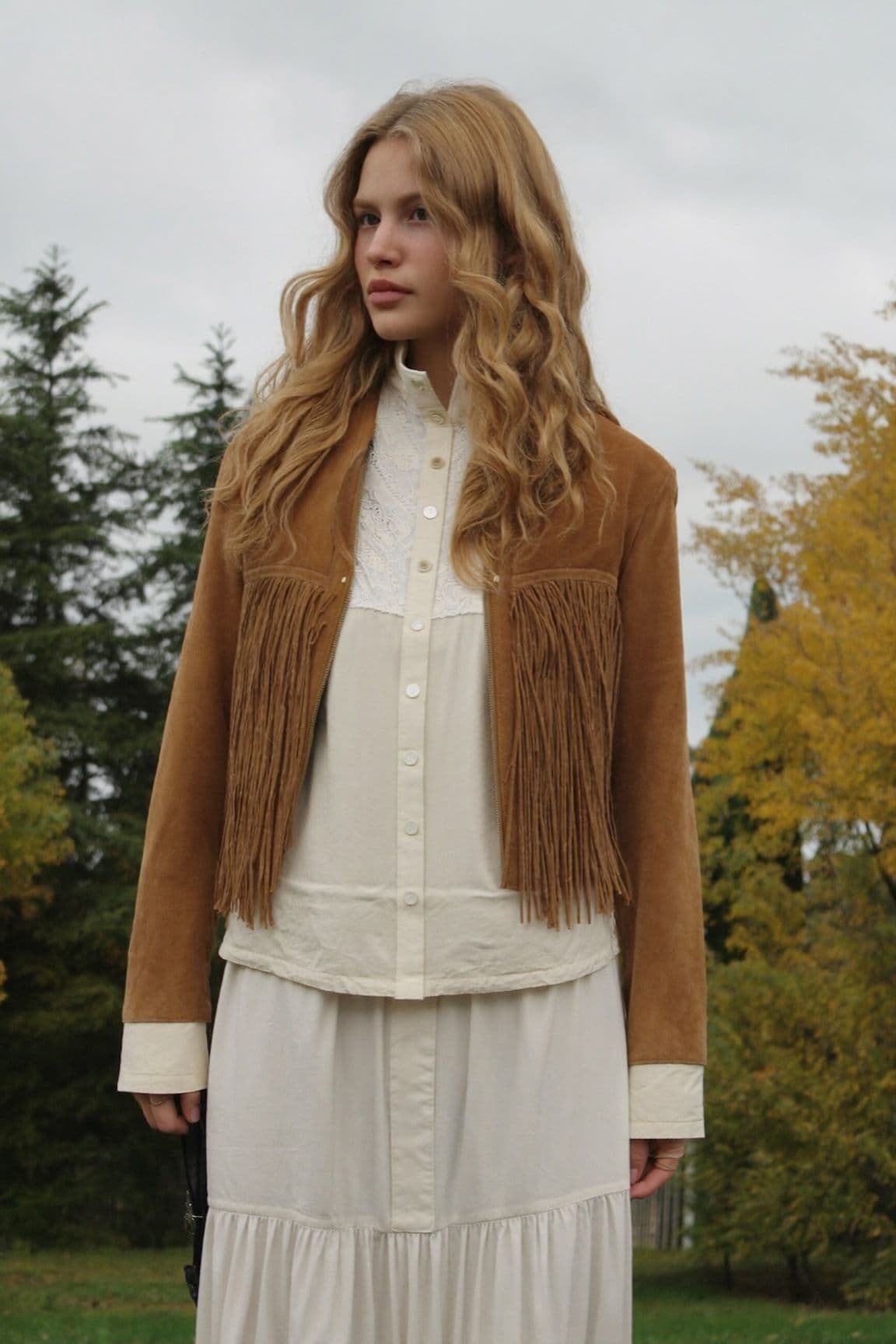 A woman with long wavy blonde hair in a tan suede fringe jacket over a cream blouse and tiered maxi skirt