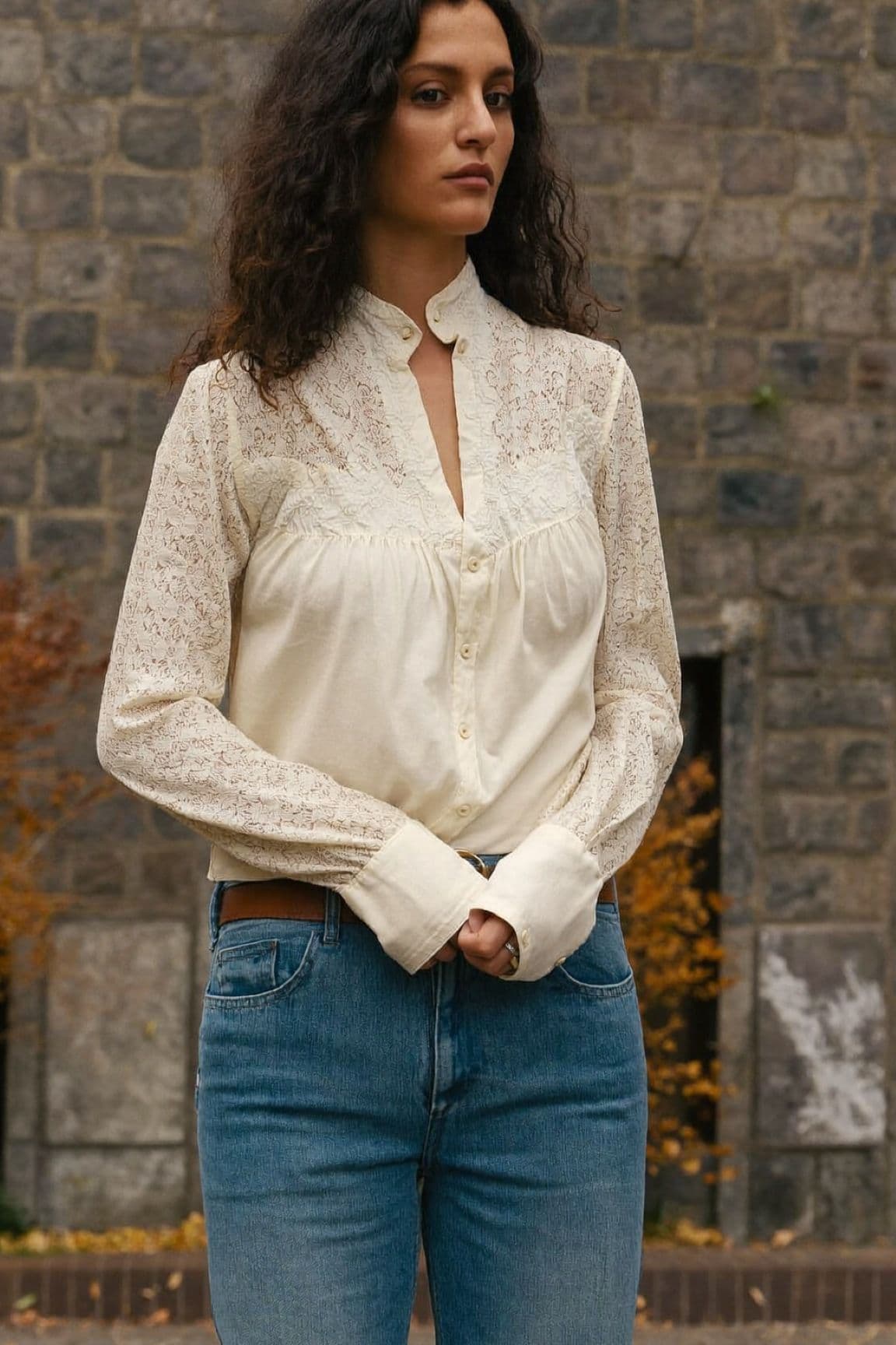 A woman with long curly hair in a cream lace blouse tucked into blue jeans in front of a stone wall
