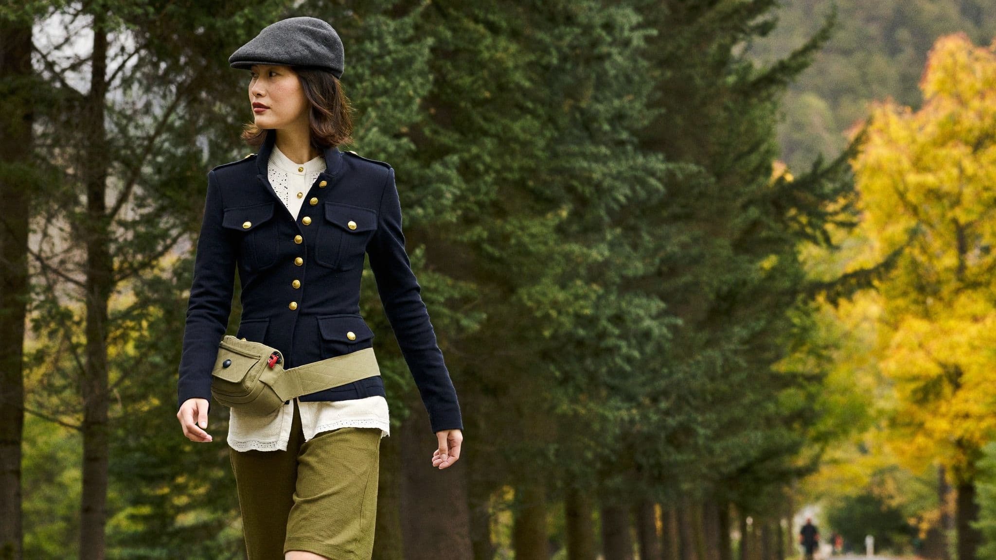 A woman walking along a tree-lined park path in a grey flat cap, a fitted navy military-style jacket with brass buttons over a cream lace-trim blouse, olive culottes and a khaki belt bag