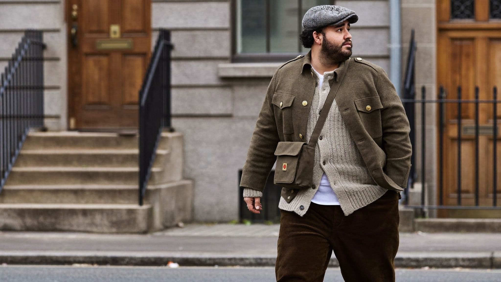 A man crossing a street of stone townhouses in a herringbone flat cap, an olive military-style jacket over a chunky oatmeal cardigan, brown corduroy trousers and a small canvas crossbody bag