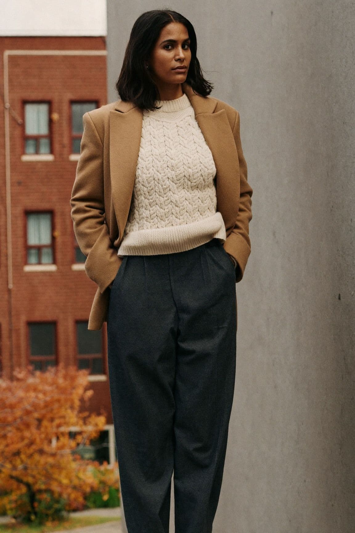 Woman standing on a Toronto sidewalk in a camel blazer over a cream cable-knit sweater with charcoal wool trousers, red brick houses and autumn trees behind her.