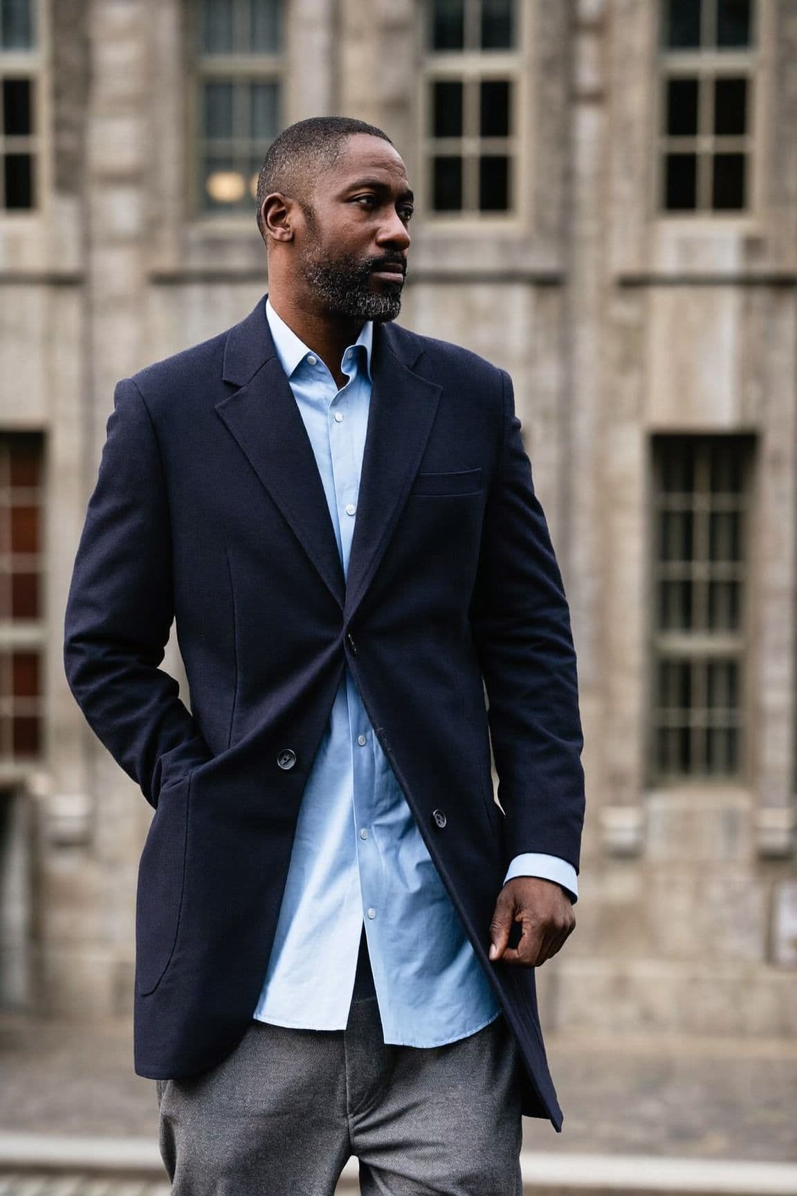 Man in a navy wool blazer over a light blue oxford shirt and grey trousers, standing in front of a grey stone building.