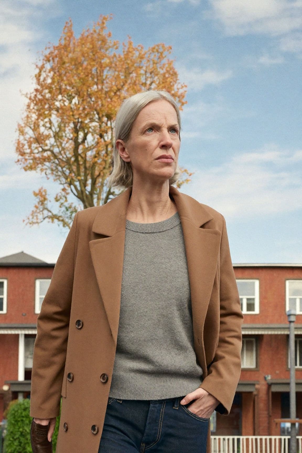 A woman with silver hair in a camel wool coat, grey crew-neck sweater and dark jeans on a street of brick row houses