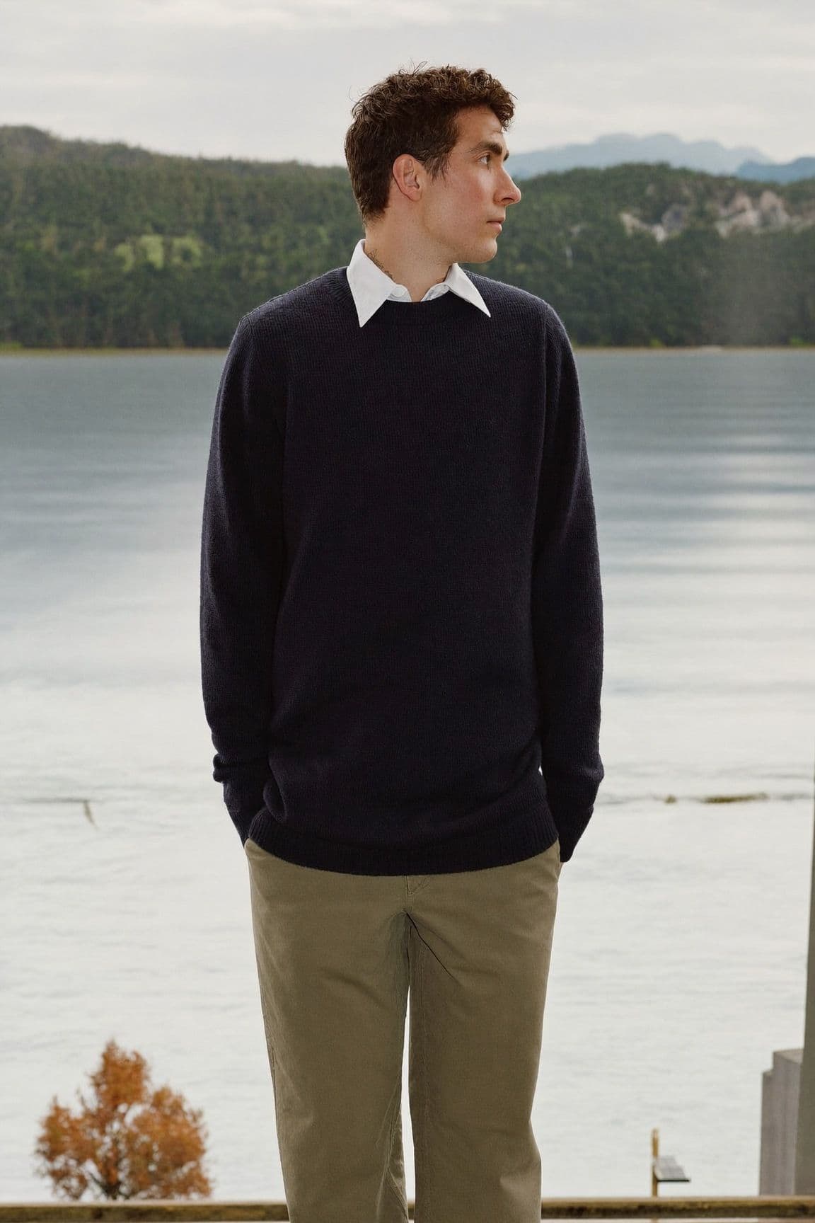 A man in a navy crew-neck sweater over a white collared shirt and olive chinos stands by the water