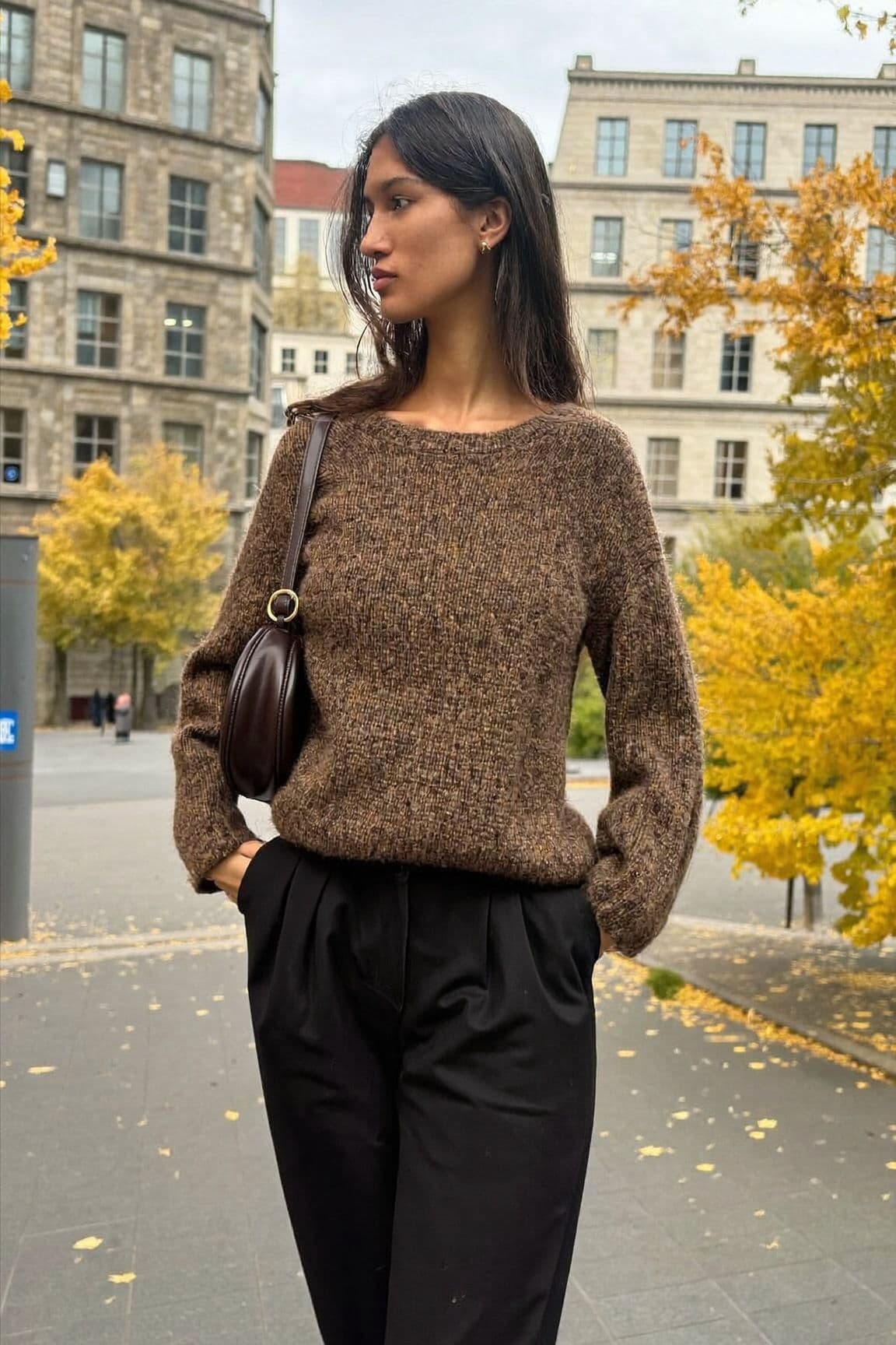 A woman in a brown chunky knit sweater and high-waisted black trousers with a small brown shoulder bag in a stone-paved square