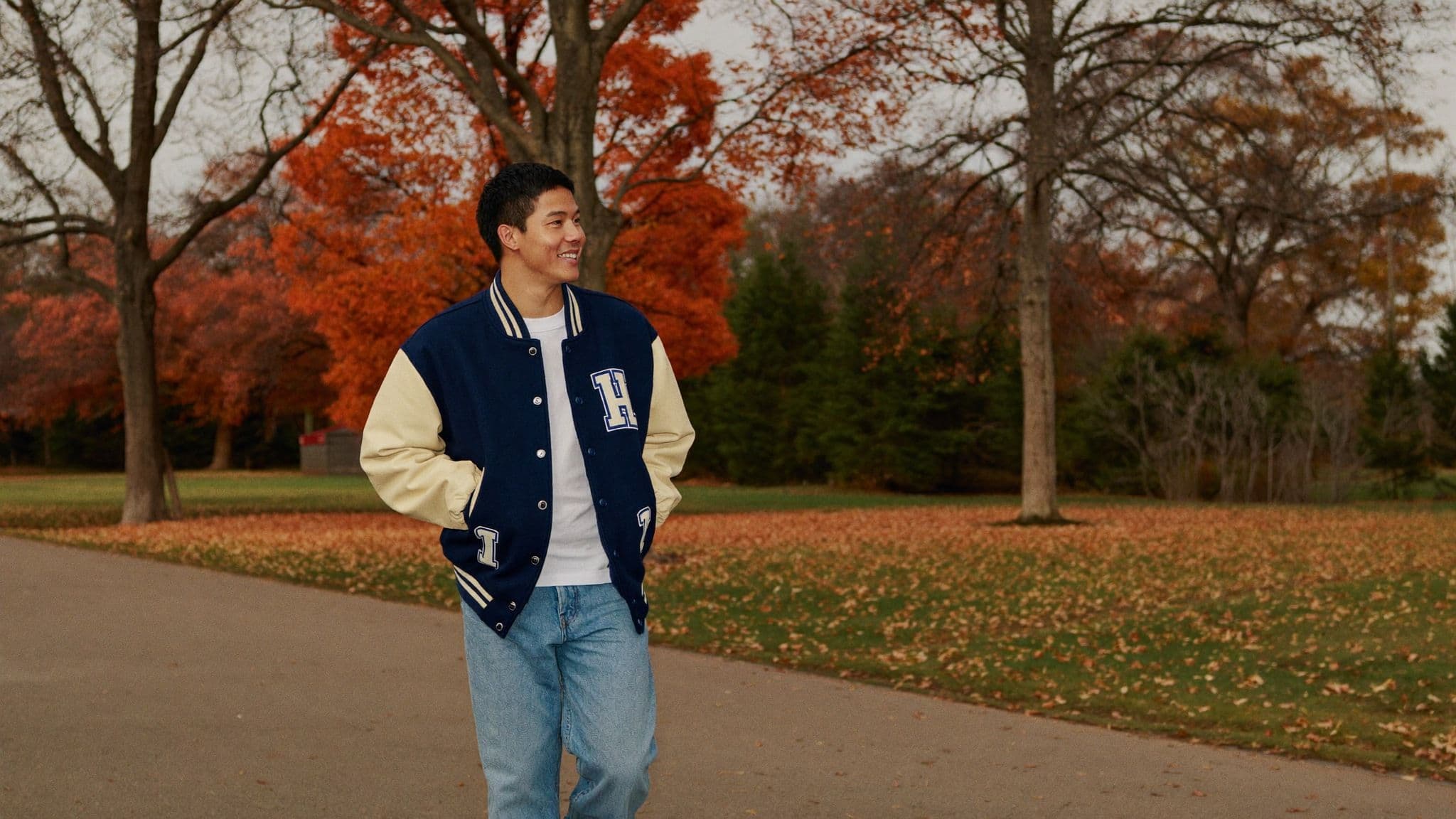 A man in a navy varsity jacket with cream sleeves, white t-shirt and light-wash jeans walks along a park path lined with autumn trees