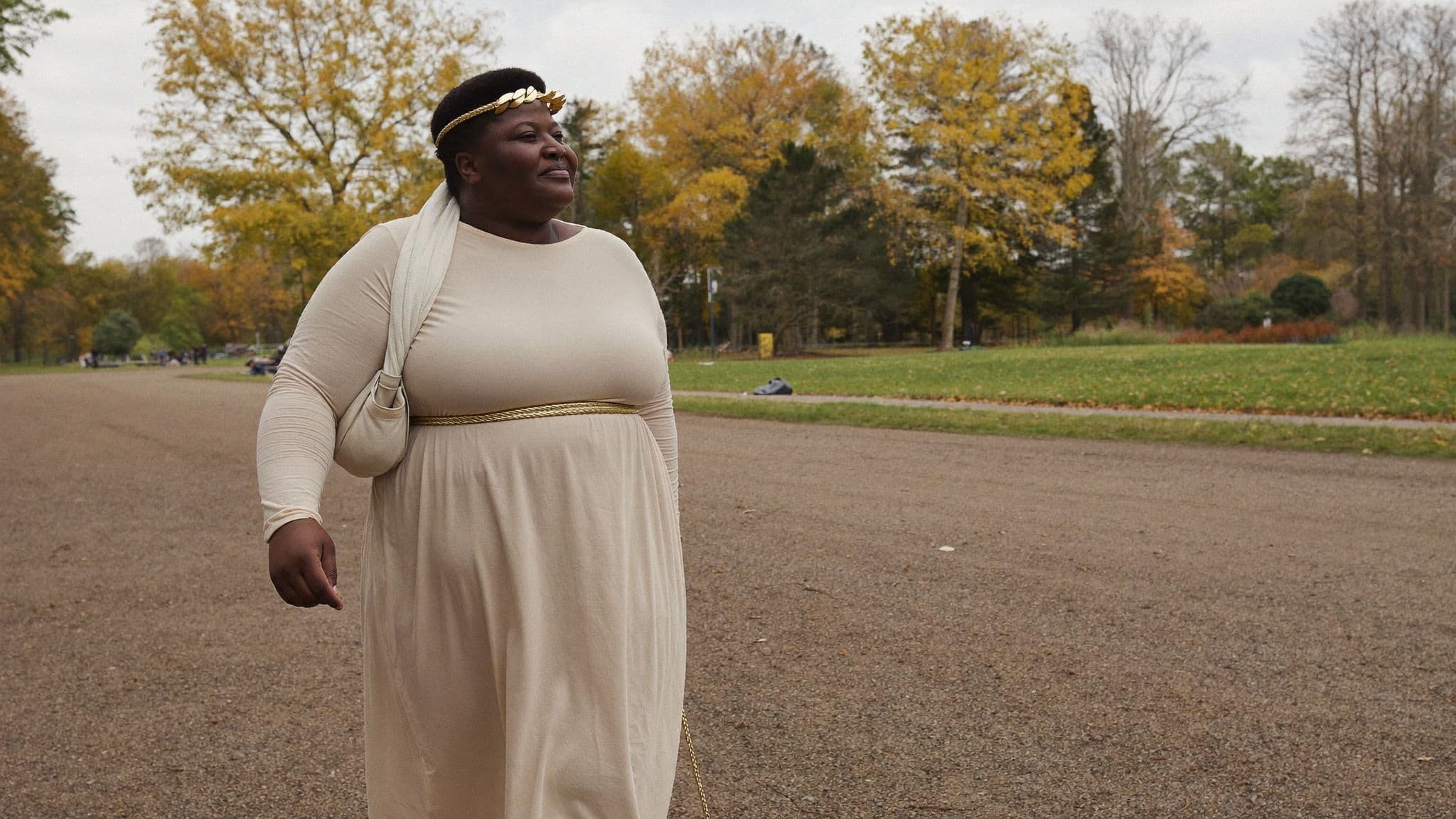 A woman in a long-sleeved cream draped dress with a gold rope belt and a gold leaf headband walks through a park in autumn
