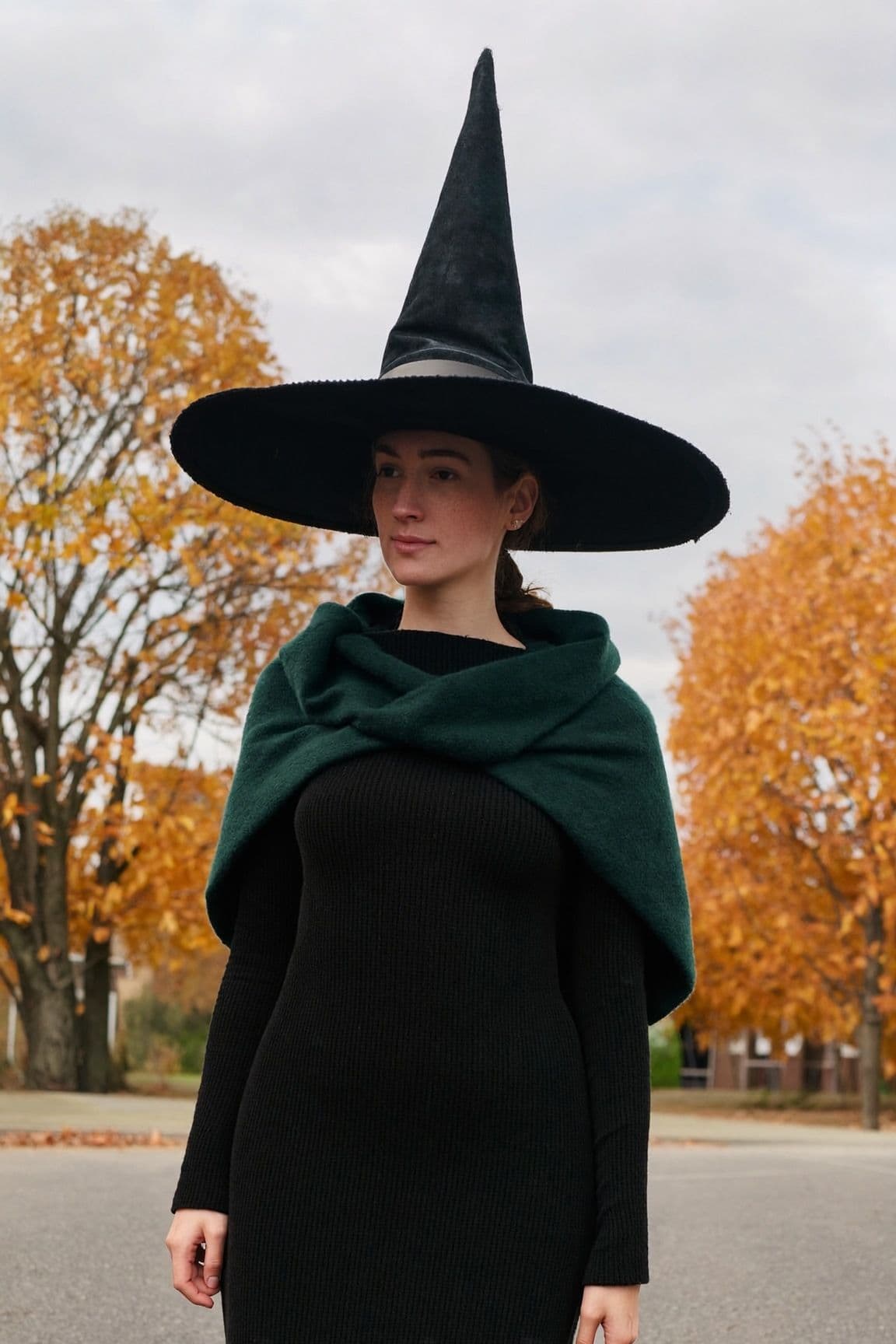 A woman in a black pointed witch hat, a black knit midi dress and a dark green shawl stands on an autumn street