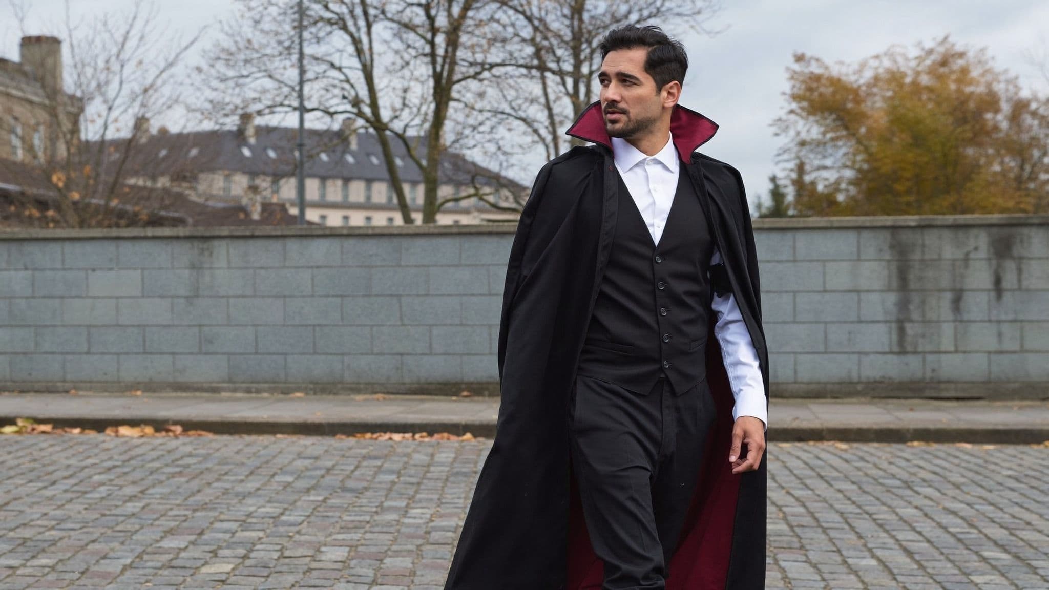 A man in a long black vampire cape with a red-lined high collar, white shirt and black waistcoat walks along a cobblestone street