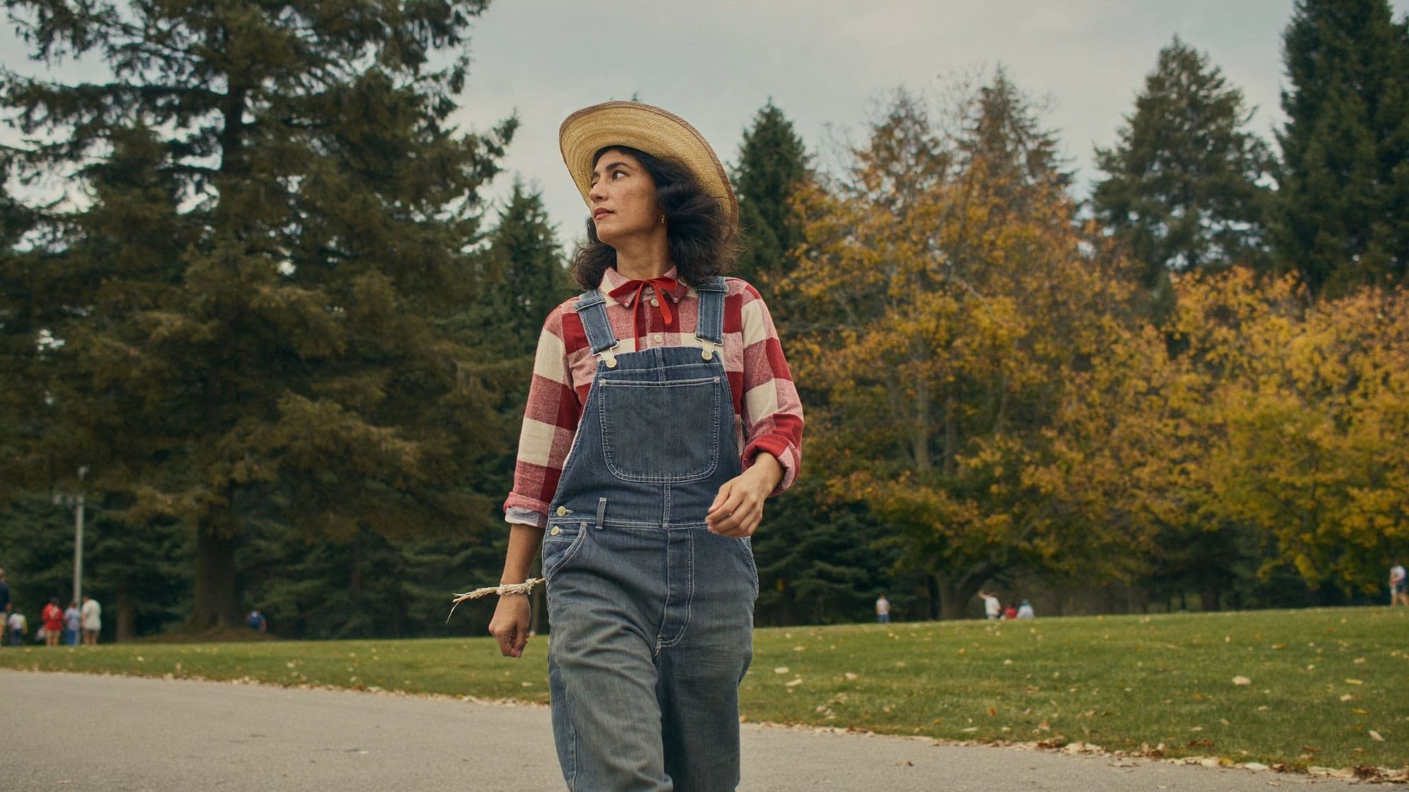 A woman in a plaid flannel shirt, denim overalls and a straw hat walks through a park in autumn