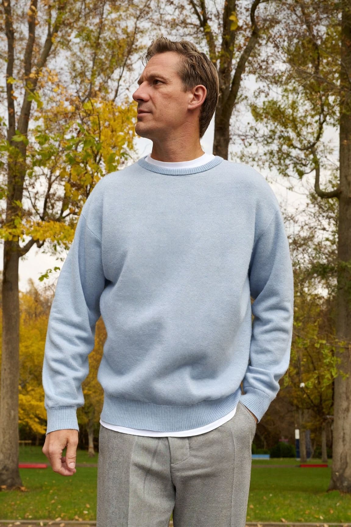 A man in a pale icy-blue crew-neck sweater and light grey trousers in a park in autumn