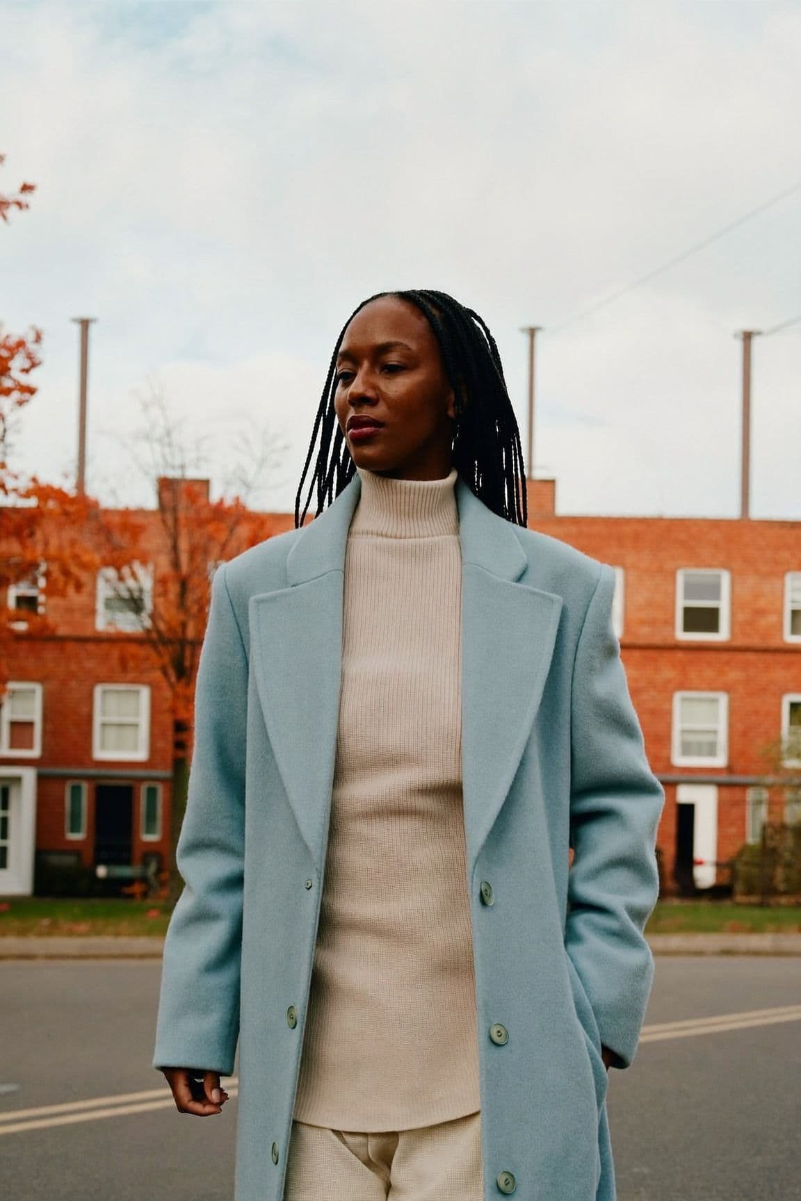 A woman with braids in a pale icy-blue wool coat over a cream turtleneck on a street of brick houses