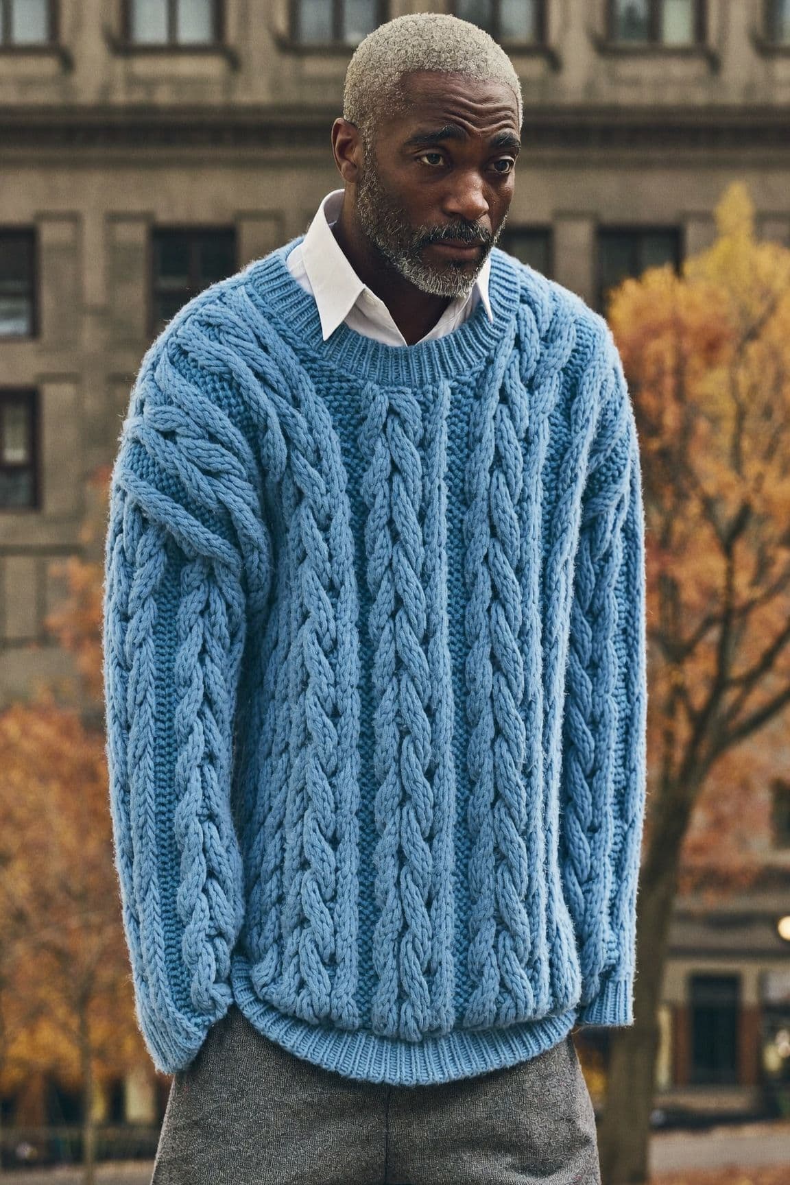 Older man with a grey beard on an autumn street wearing an oversized pale blue cable-knit sweater over a white collared shirt with grey trousers