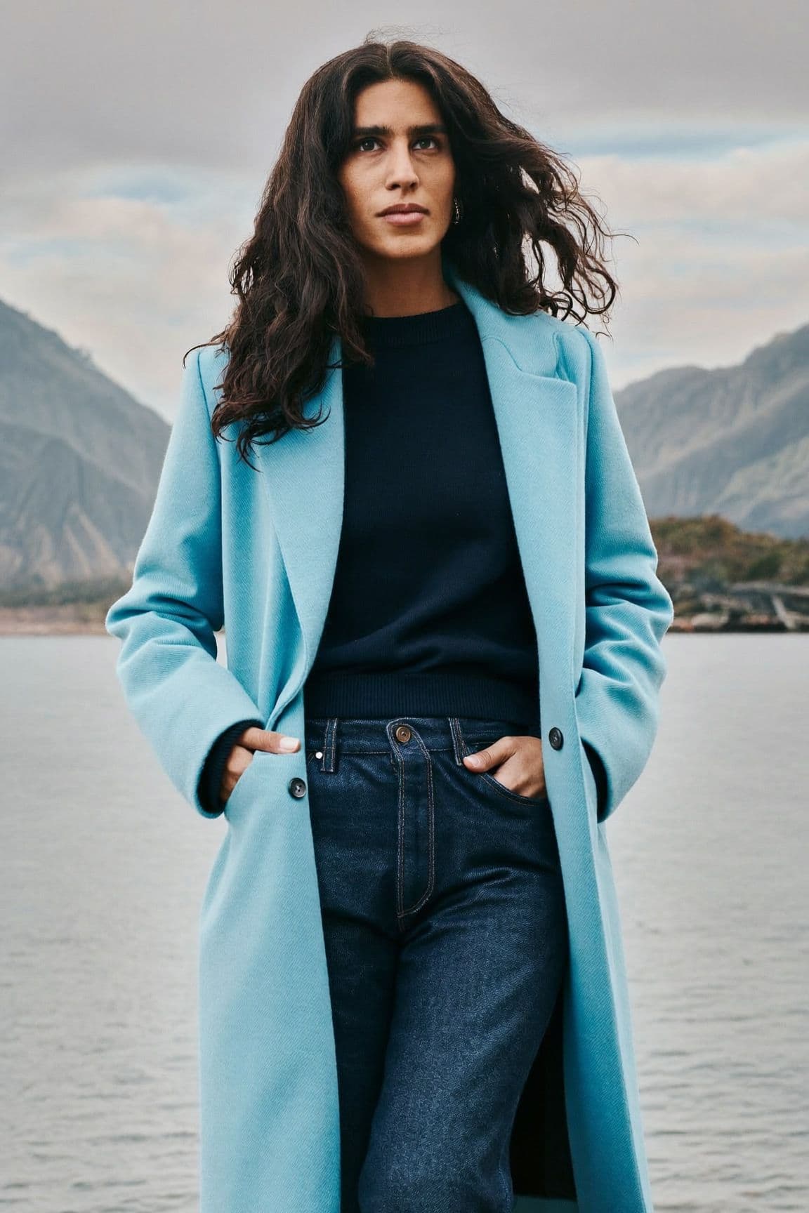 Woman by the water with mountains behind her wearing a long icy blue wool coat open over a navy sweater and dark straight-leg jeans