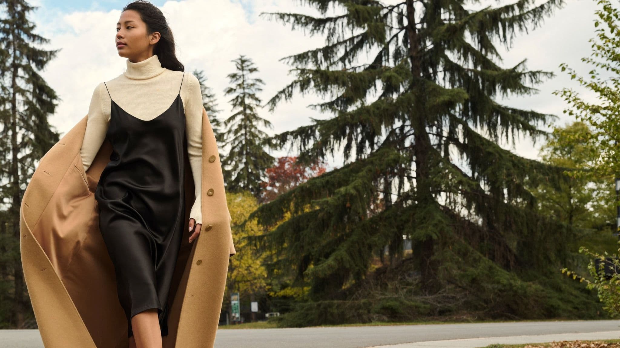 Woman walking among evergreen trees in Vancouver wearing a cream ribbed turtleneck under a black satin slip dress, with a long camel duster coat worn open