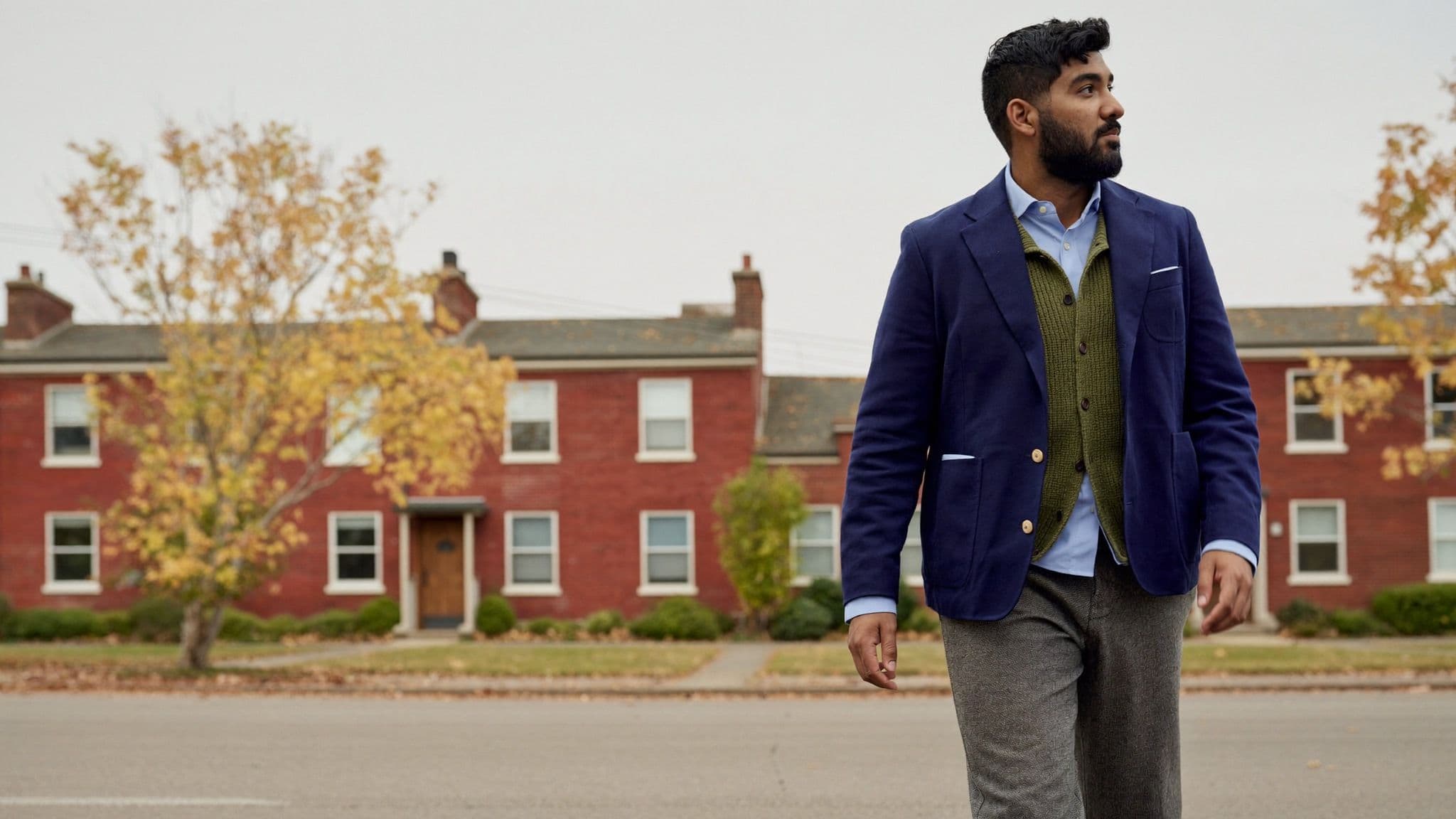 Man walking down a Toronto street of red brick houses in a navy blazer over an olive knit layer and a pale blue oxford shirt, with grey wool trousers
