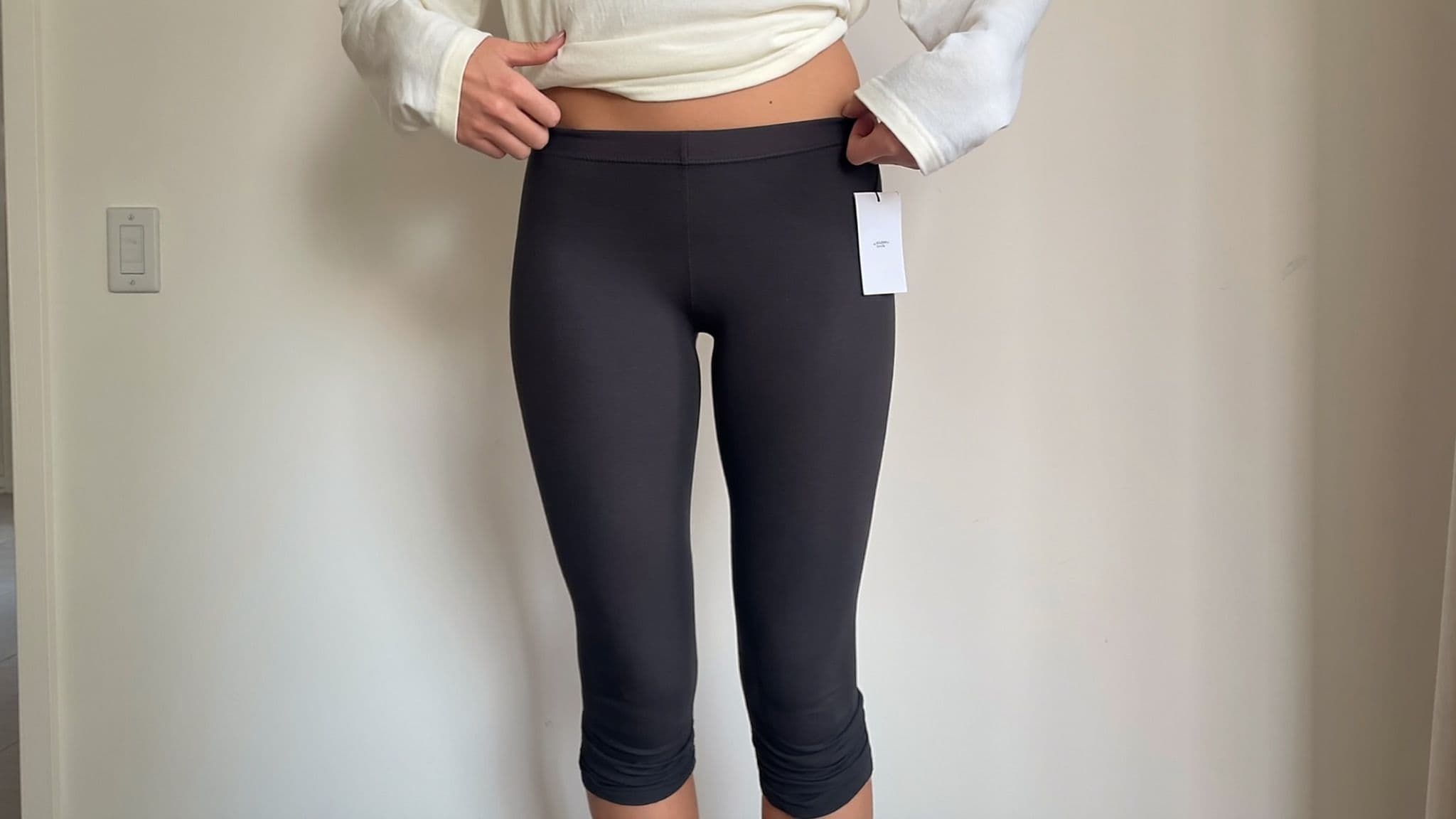 Woman in black leggings and a cream top adjusting the leggings waistband with both hands