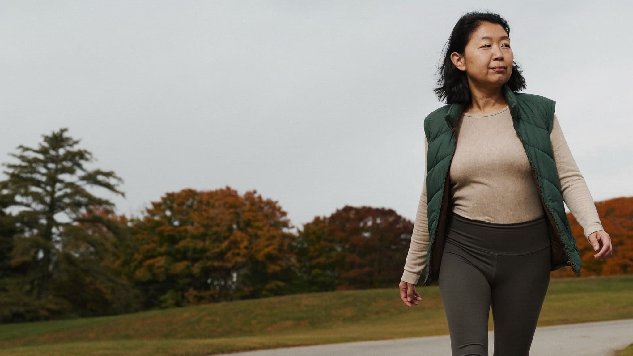 Woman in charcoal high-rise leggings, an oatmeal ribbed top and a green quilted vest walking in a park in autumn