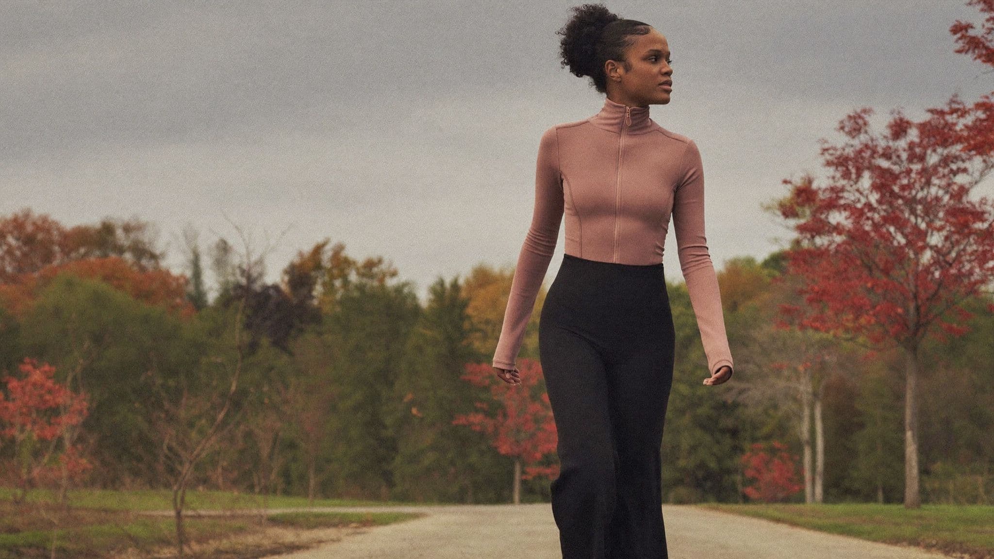 Woman in a fitted dusty-rose zip jacket and black flared leggings walking on a park path with red autumn trees