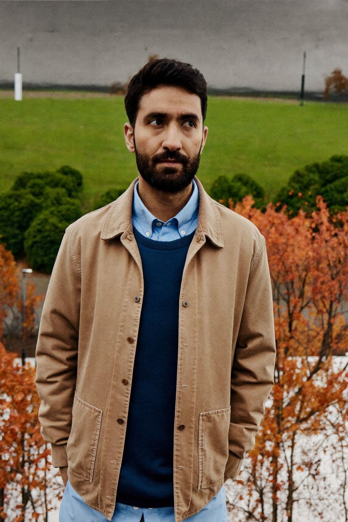 A man in a light-blue oxford shirt under a navy sweater and an open tan chore jacket in a park in autumn