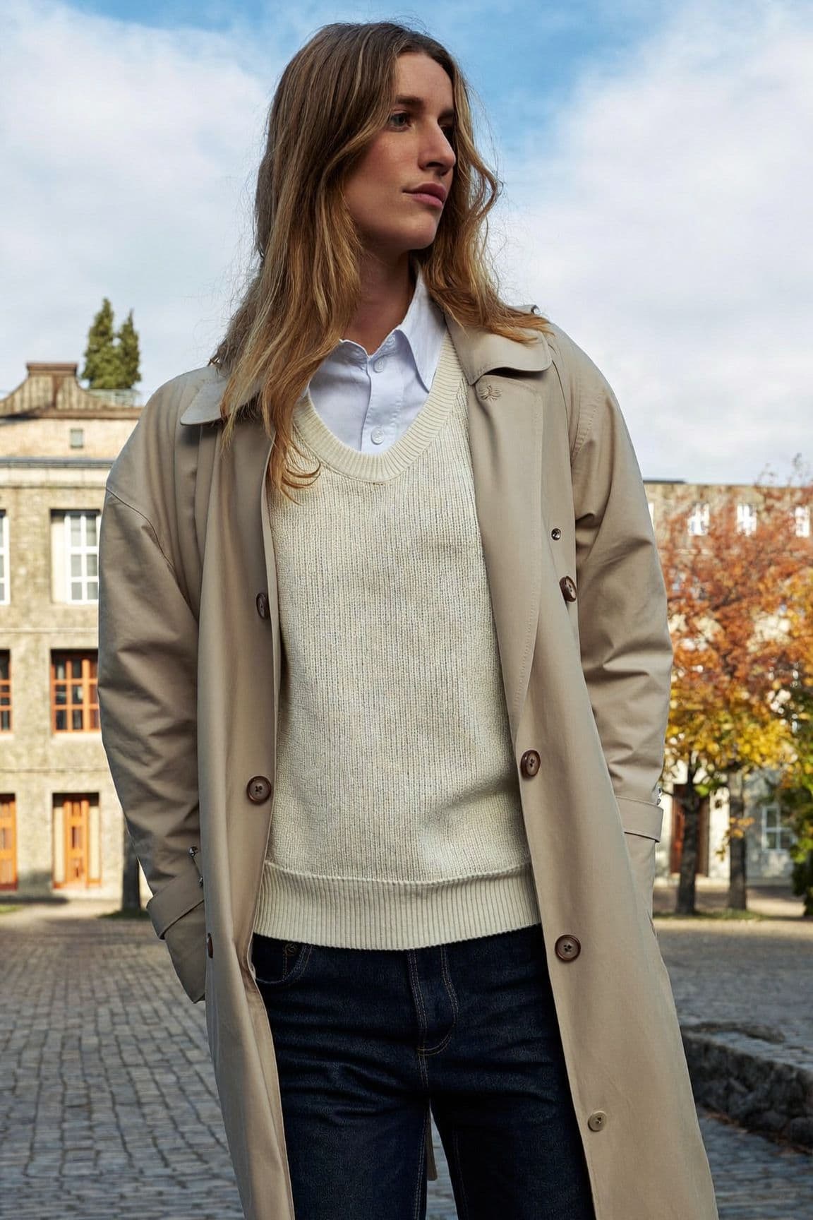 A woman in a cream knit vest over a white shirt under a beige trench coat with dark jeans on a cobblestone street