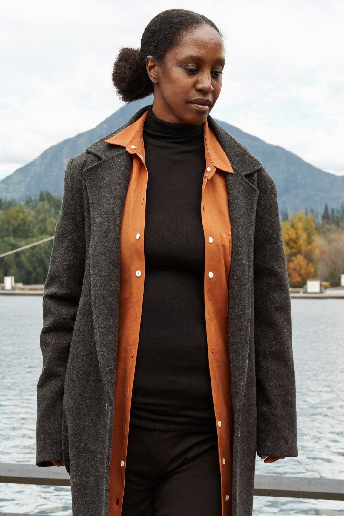 A woman in a black turtleneck under an open rust shirt jacket and a long charcoal wool coat by the water