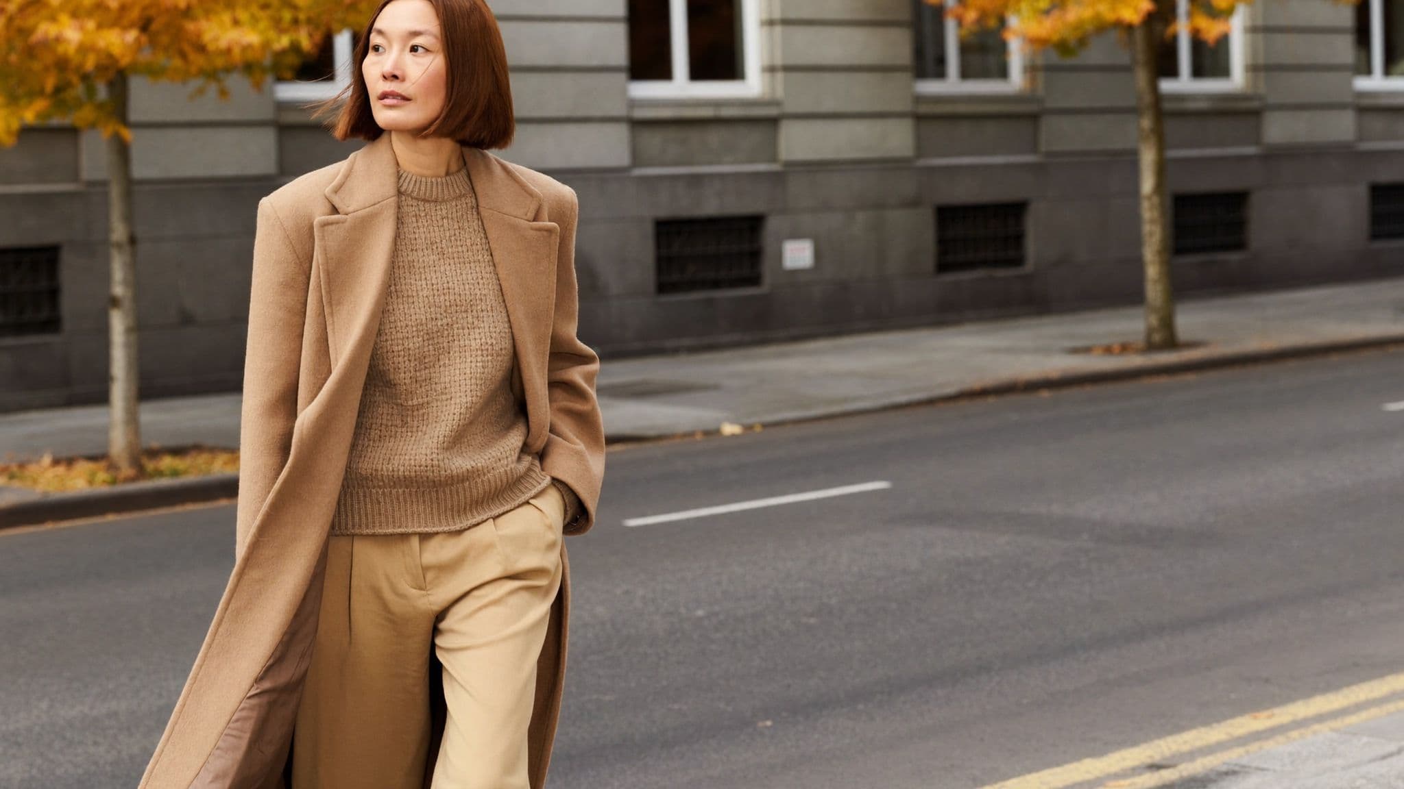 Woman walking down an autumn street wearing a long camel wool coat open over a textured oatmeal knit sweater and sand-coloured wide-leg trousers