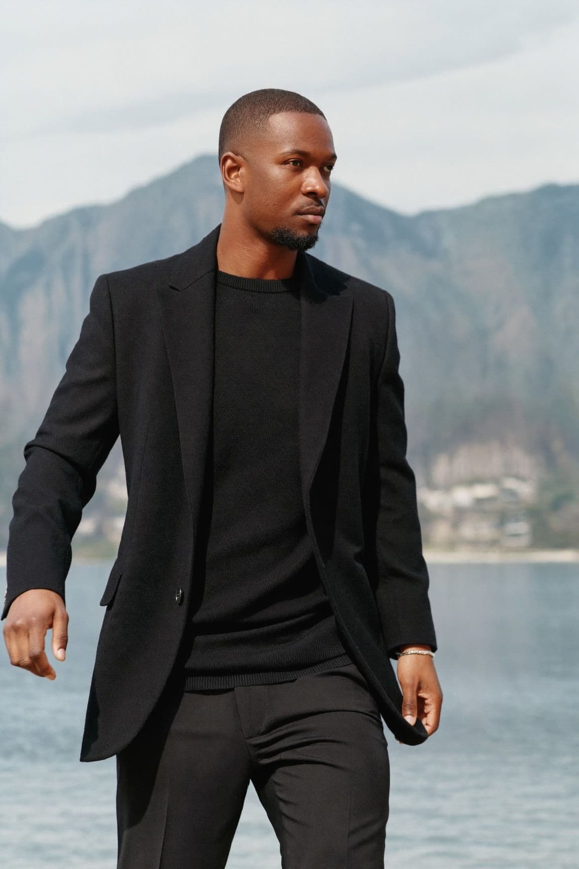 Man walking by the water with mountains behind him, wearing a relaxed black blazer over a fine black knit with black trousers and a thin silver bracelet