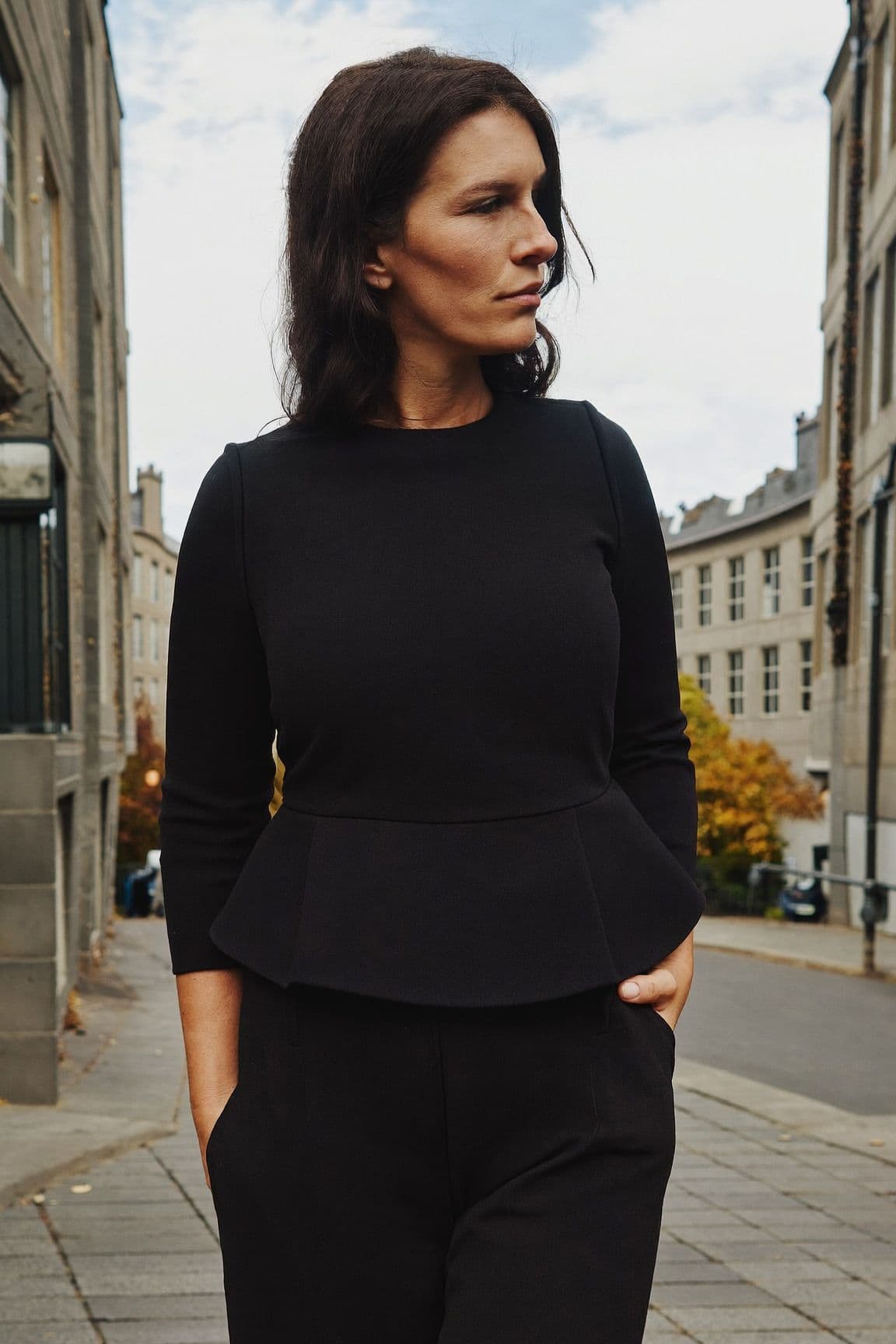 A woman in a structured black peplum top with black wide-leg trousers on a stone-paved street