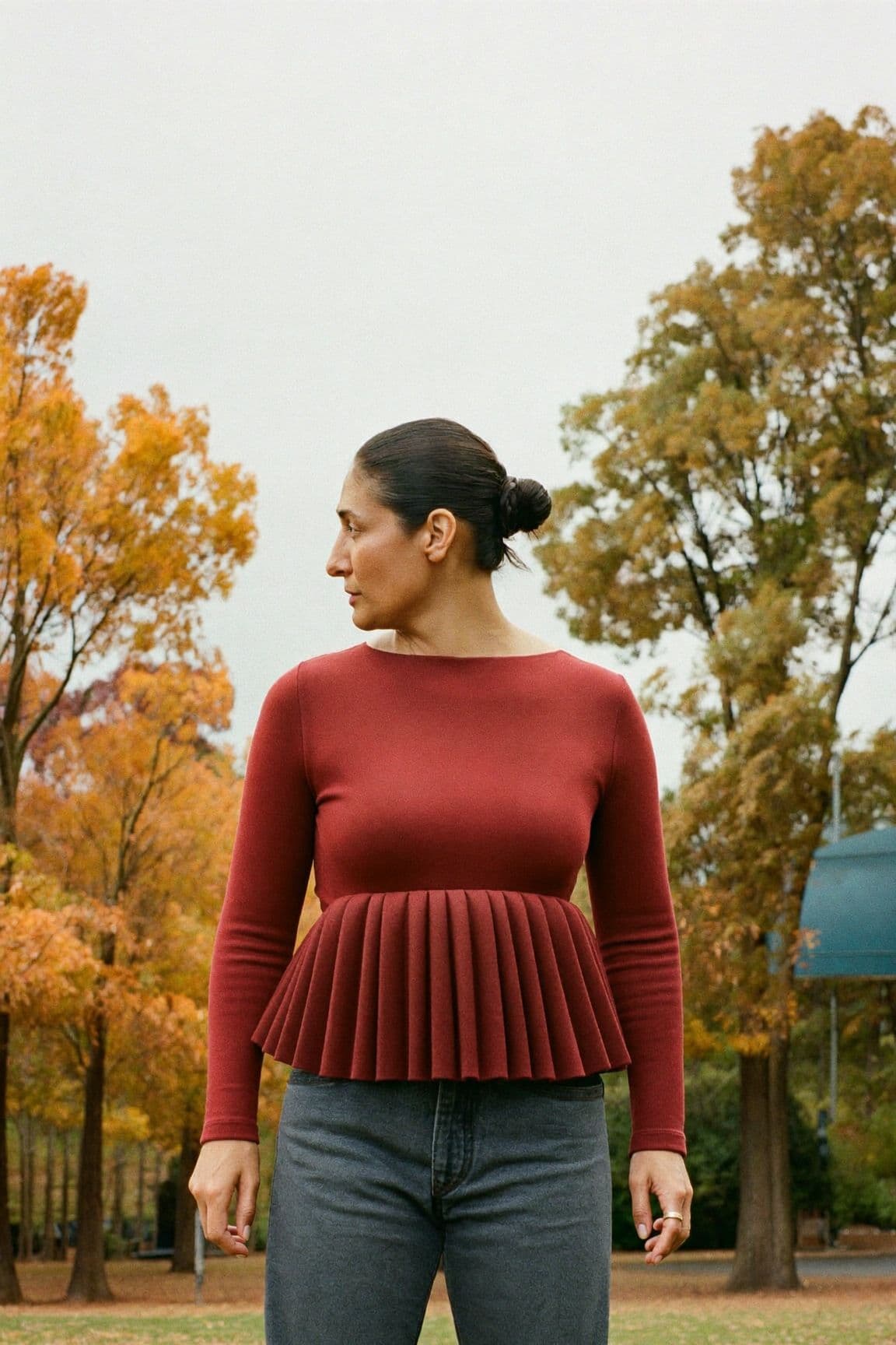 A woman in a burgundy long-sleeved top with a pleated peplum at the waist and grey jeans in a park