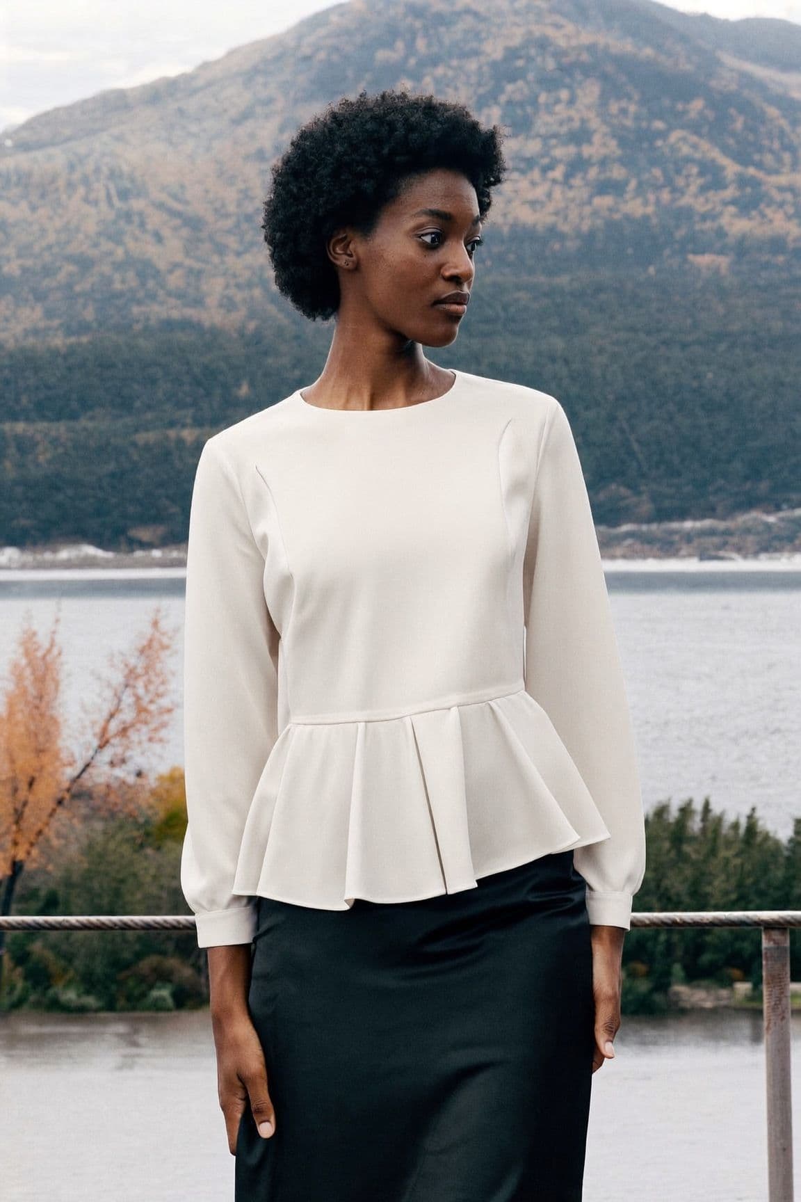 A woman in an ivory peplum blouse and a black midi skirt by the water