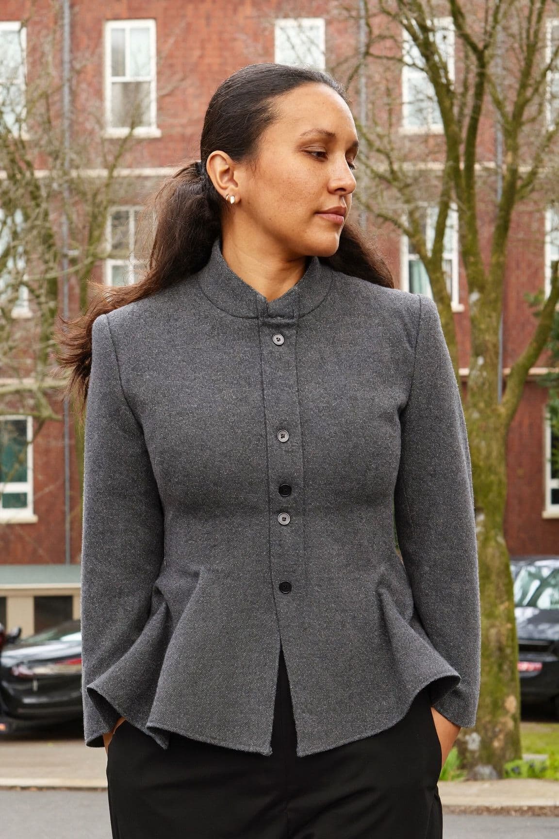 A woman with a long dark ponytail in a structured charcoal wool peplum jacket with a stand collar, buttoned up, with a short flare at the hip over black trousers