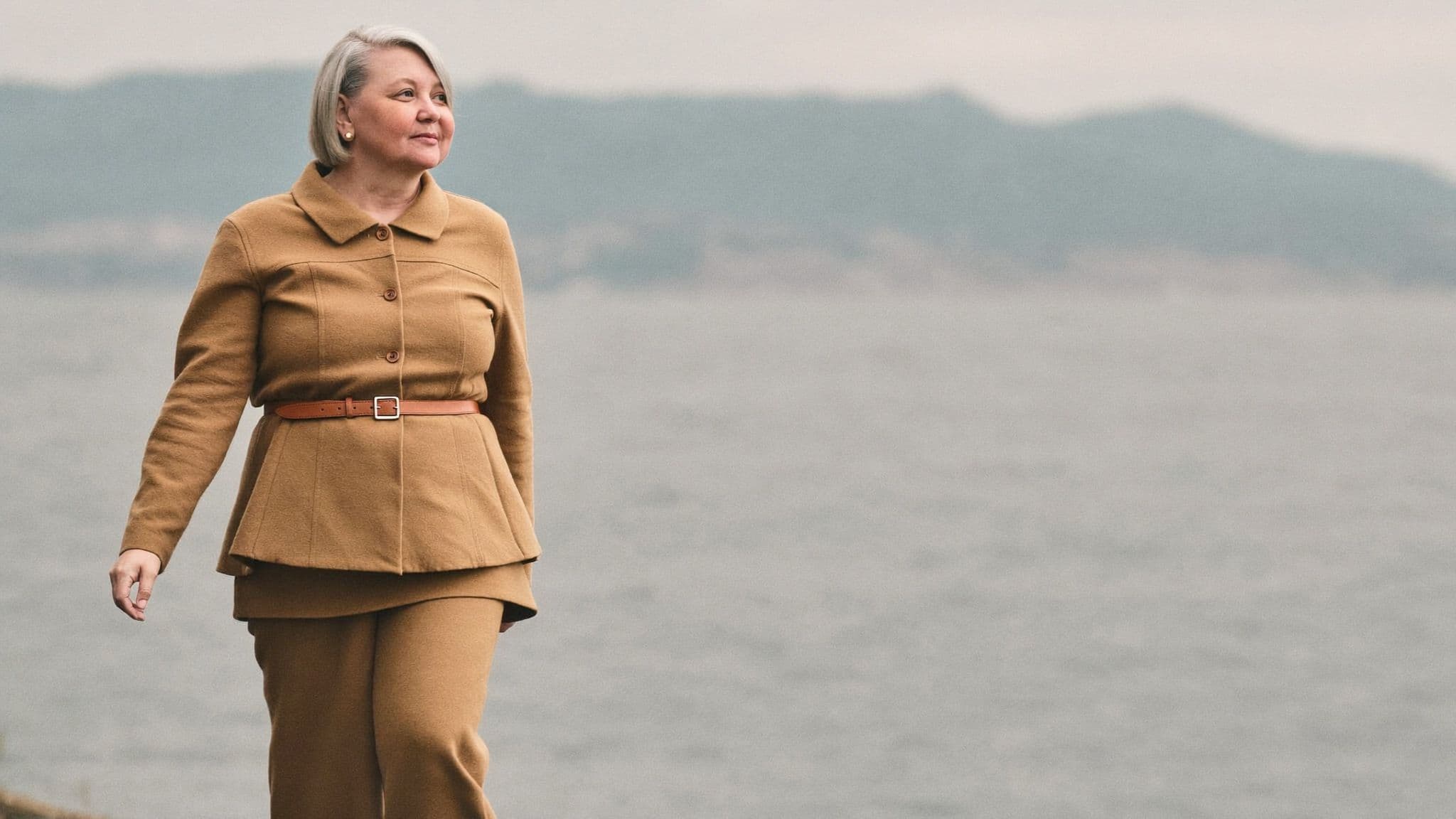 A woman with a silver bob walking by the water in a tone-on-tone camel outfit: a belted camel peplum jacket and matching straight-leg trousers