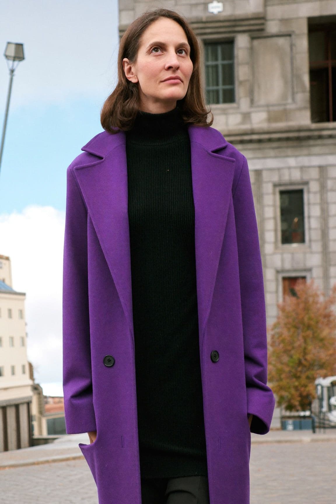 A woman in a royal purple wool coat over a black turtleneck standing in front of stone buildings