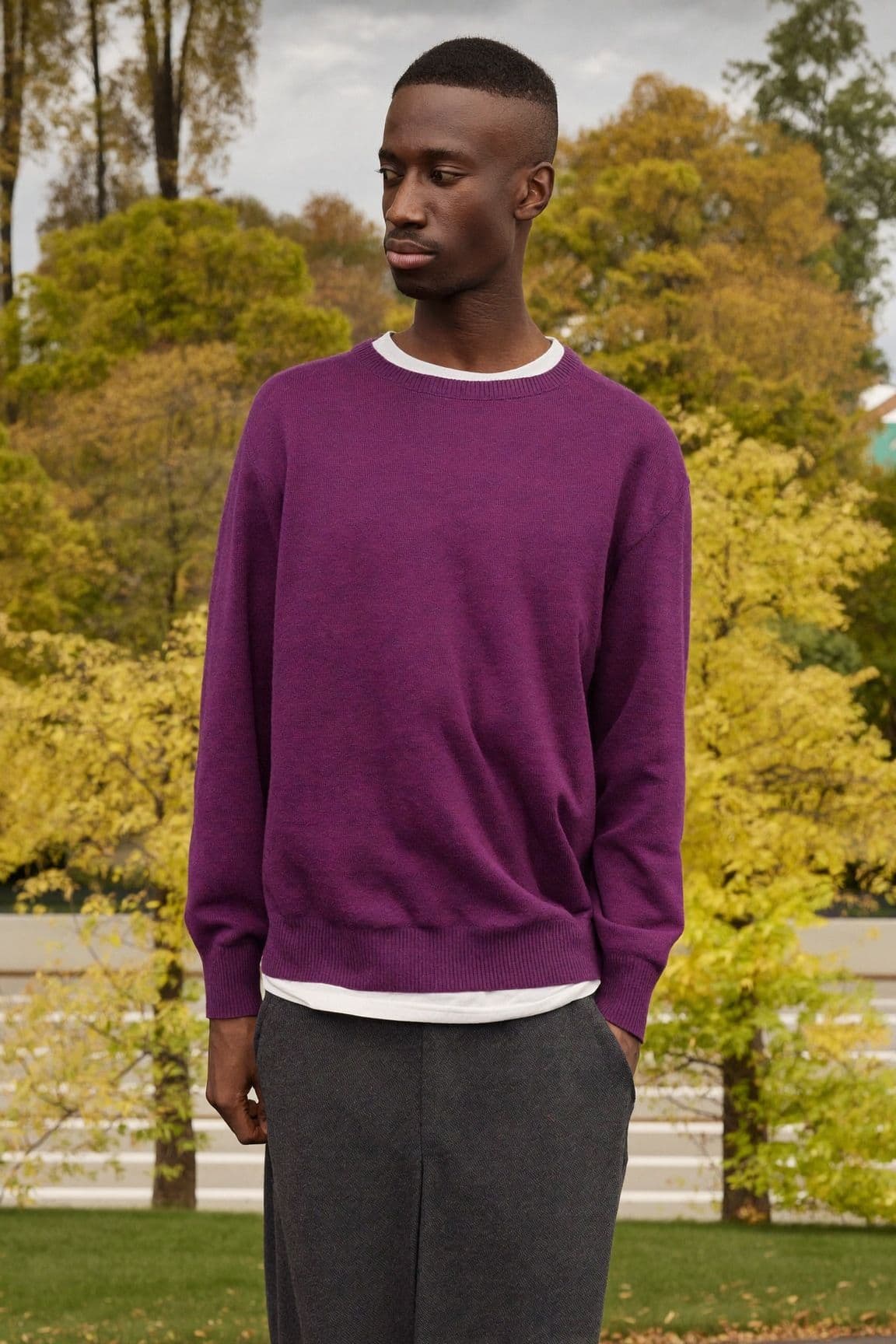 A man in a deep purple crew-neck sweater with a white t-shirt hem showing and charcoal trousers in a park