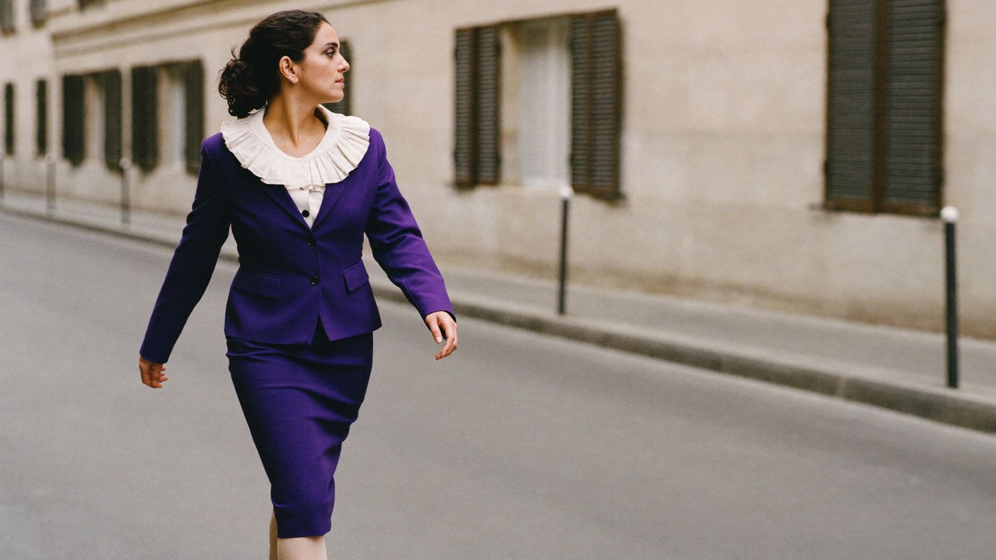 Woman walking down a Milan street in a royal purple skirt suit with a slim jacket and knee-grazing pencil skirt over an ivory ruffled-collar blouse.