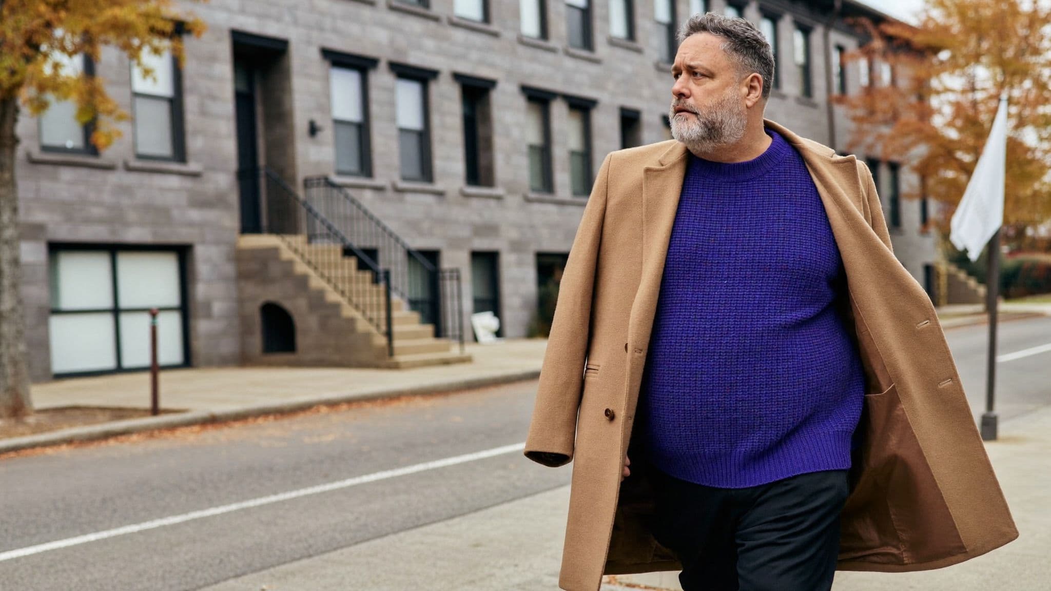 Older man with a grey beard walking on a residential street in a royal purple chunky knit sweater, black trousers and an open camel overcoat.