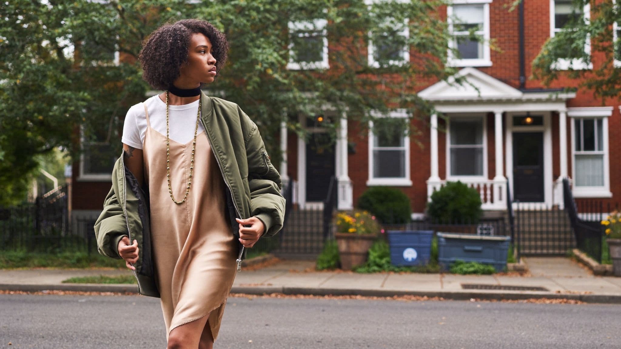 Woman walking on a residential street in a champagne slip dress layered over a white t-shirt, a black choker with a long gold chain and an olive bomber jacket.