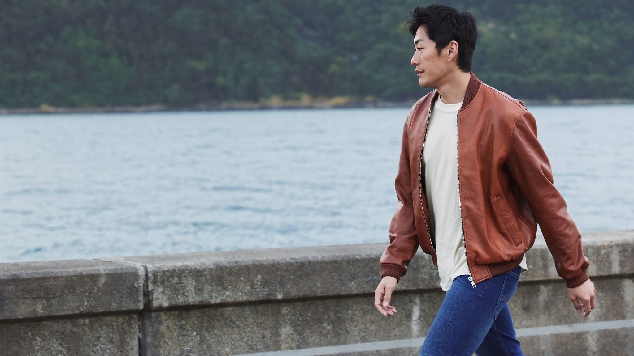 Man walking along a seawall by the water in a relaxed cognac leather bomber jacket, cream t-shirt and bright cobalt blue skinny jeans.